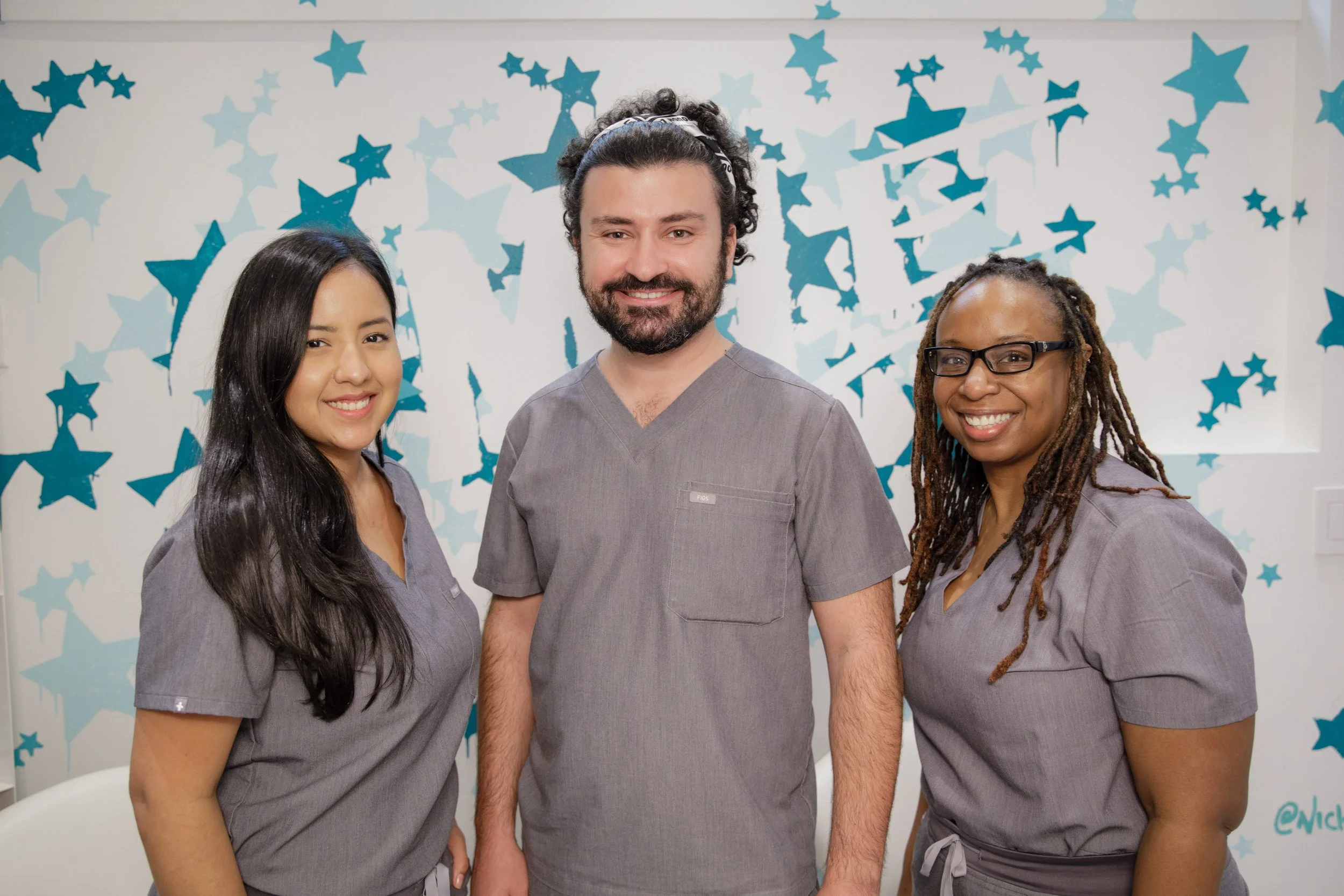 Three healthcare workers, two women and one man, smiling in front of a blue star-themed decoration.