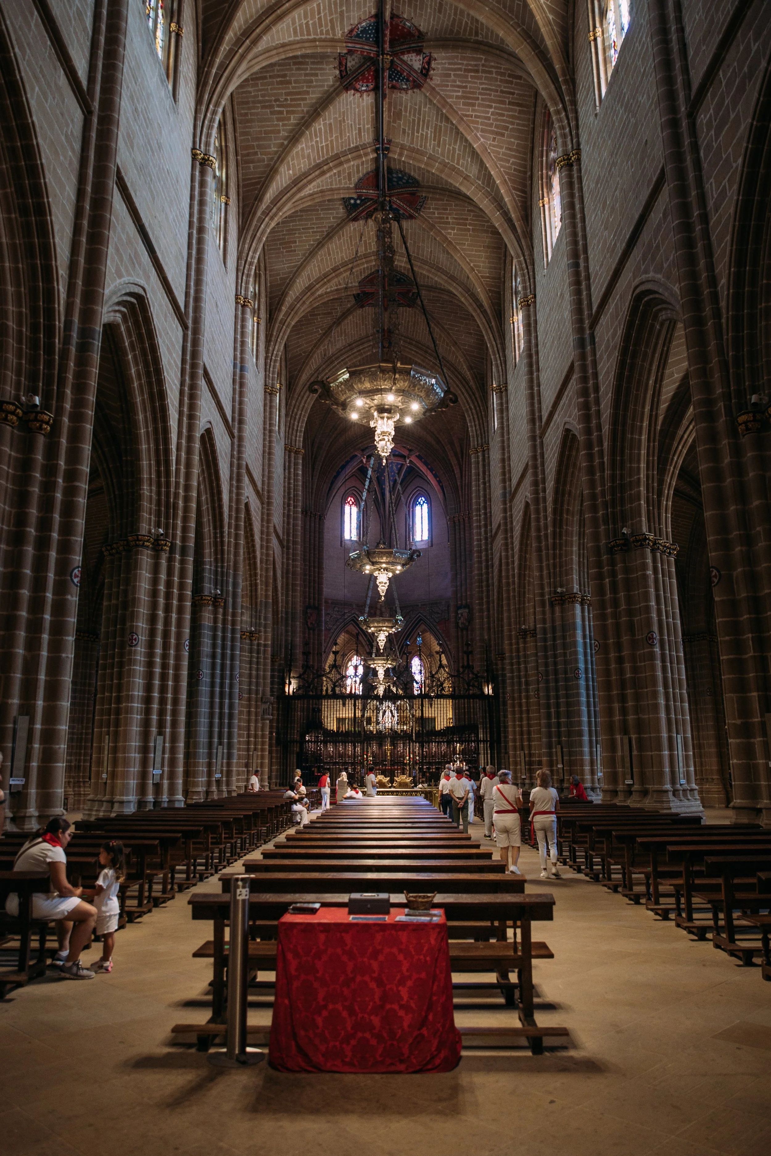 Inside of a large church with high vaulted ceilings, stained glass windows, and chandeliers. People are walking and sitting on wooden pews.