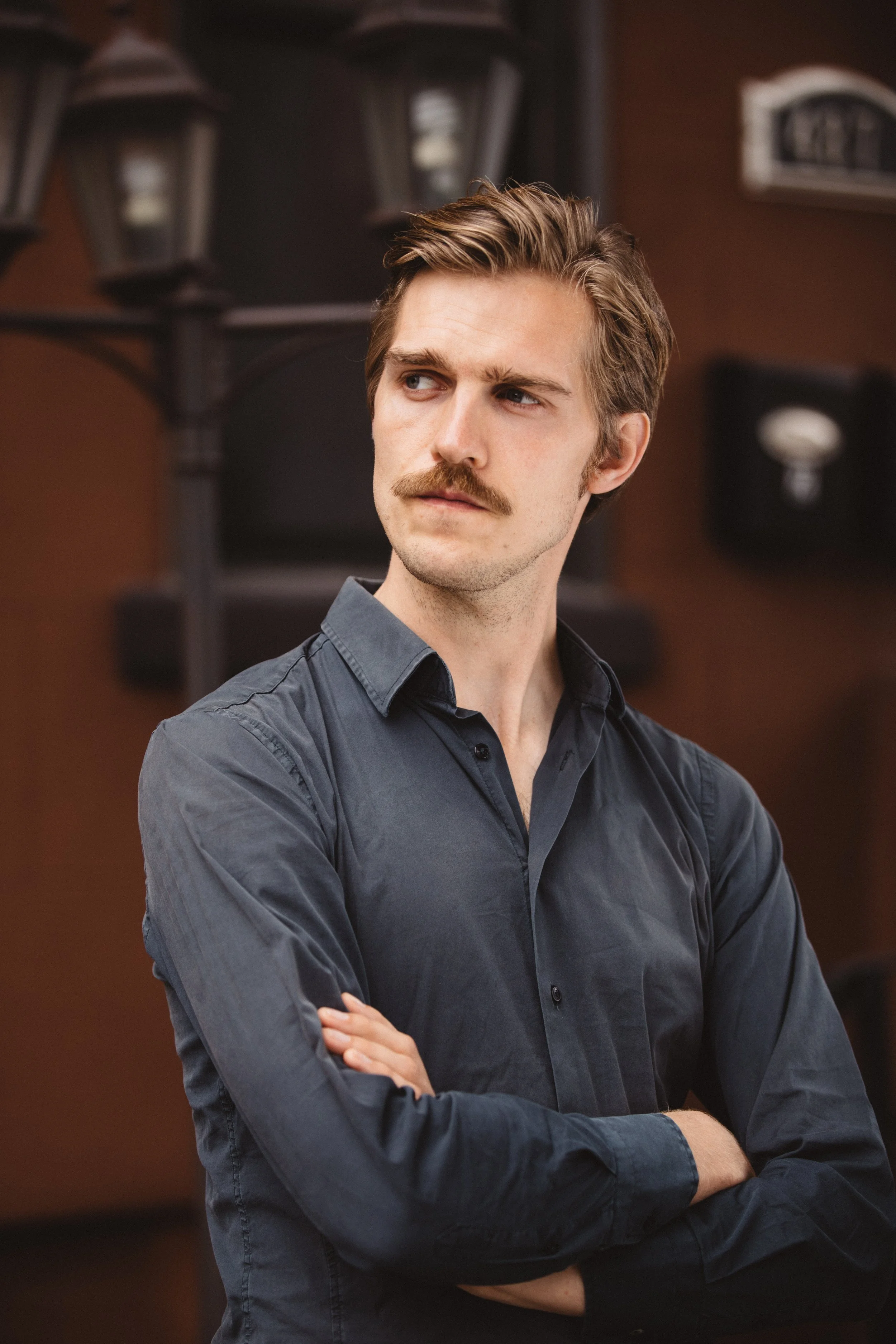 A young man with light brown hair, a mustache, and a serious expression is standing with his arms crossed, wearing a dark button-up shirt, in front of a background with outdoor lanterns and a building with the number 1822.