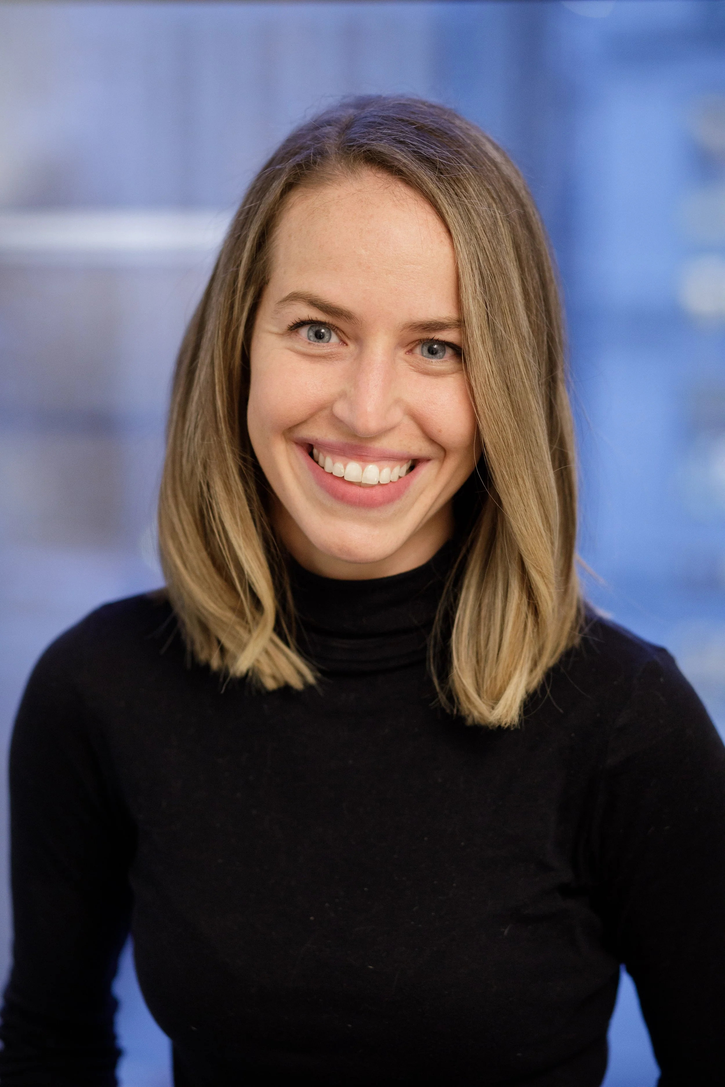 Close-up of a woman with shoulder-length blonde hair, blue eyes, smiling, wearing a black turtleneck, against a blurred blue background.