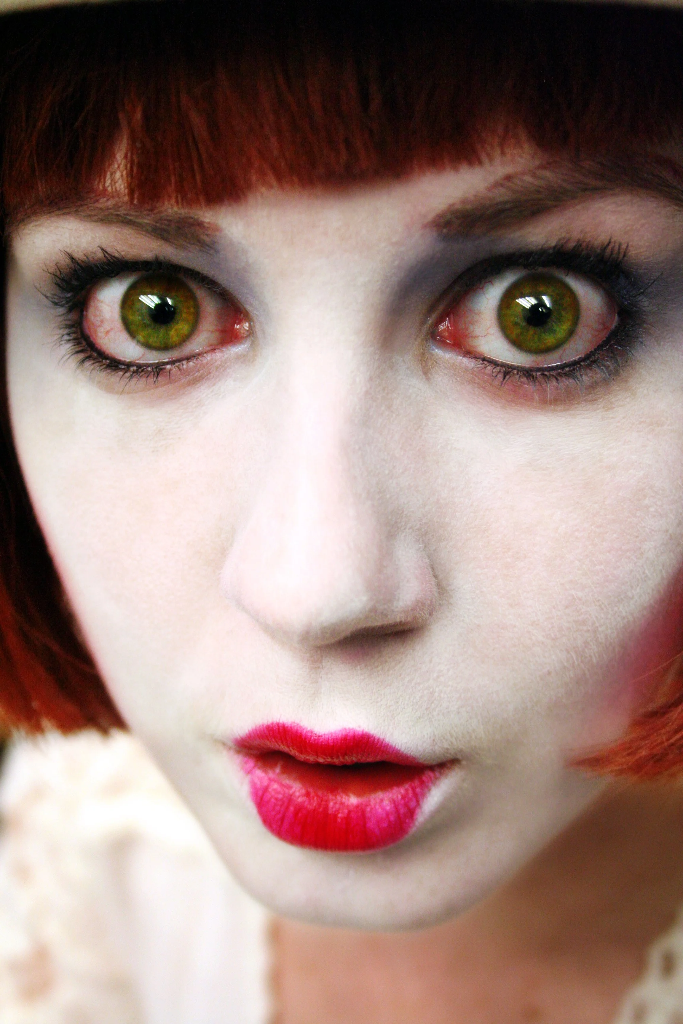 Close-up of a woman with red hair, green eyes, white makeup, and pink lipstick.