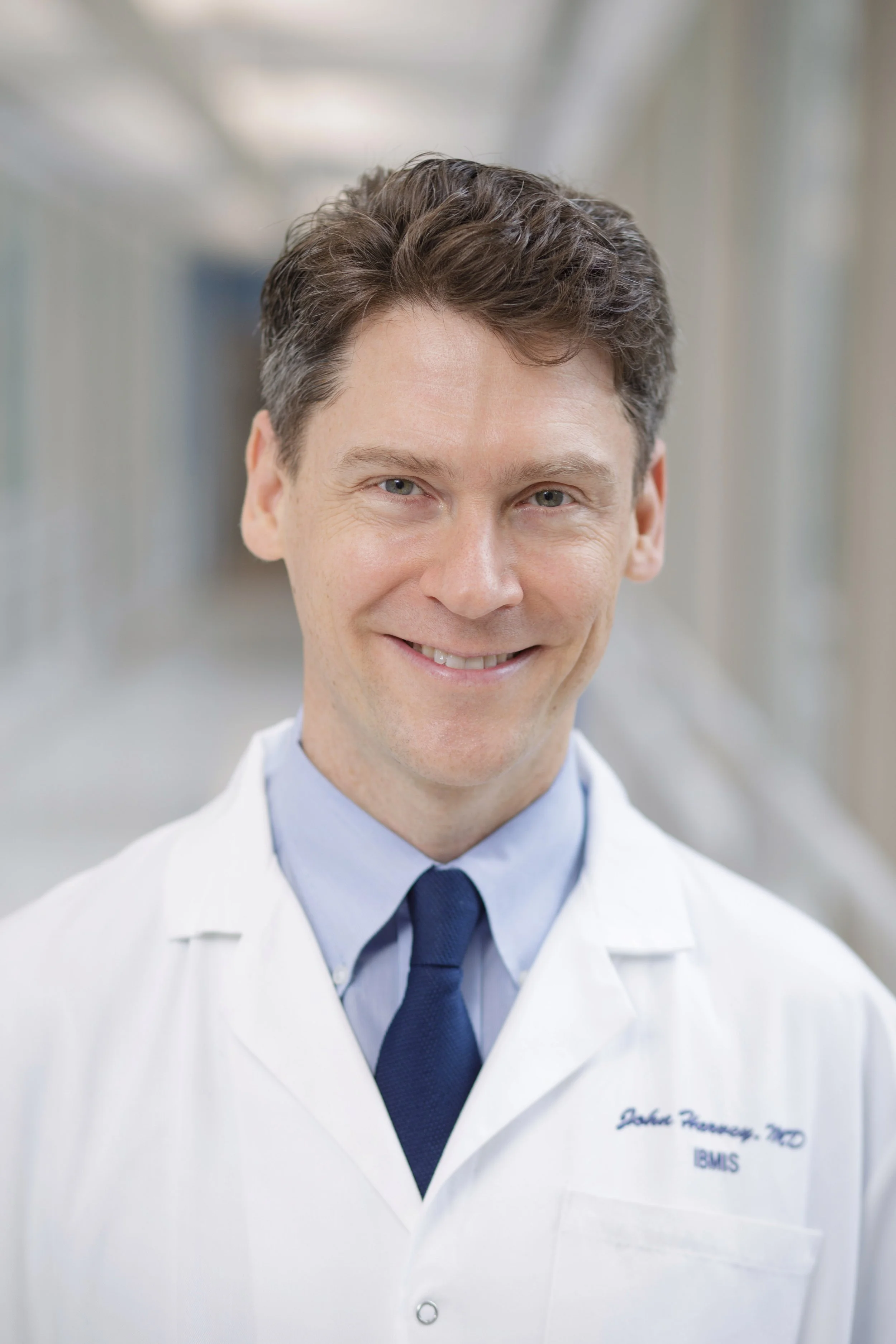 A smiling male doctor in a white lab coat with a blue tie, in a hospital corridor.