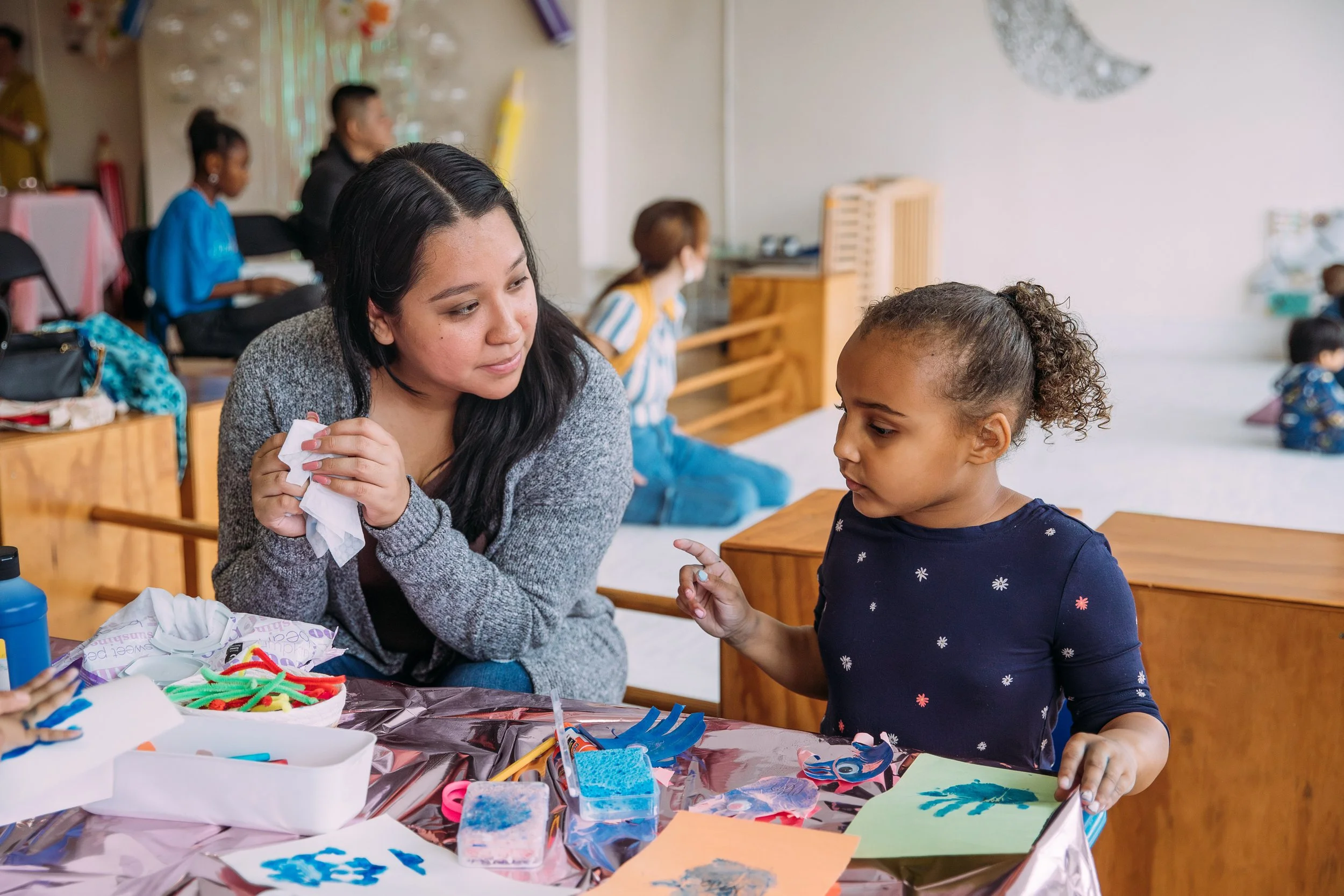 A woman and a young girl at a table decorated with arts and crafts supplies, engaged in a creative activity in a classroom setting.