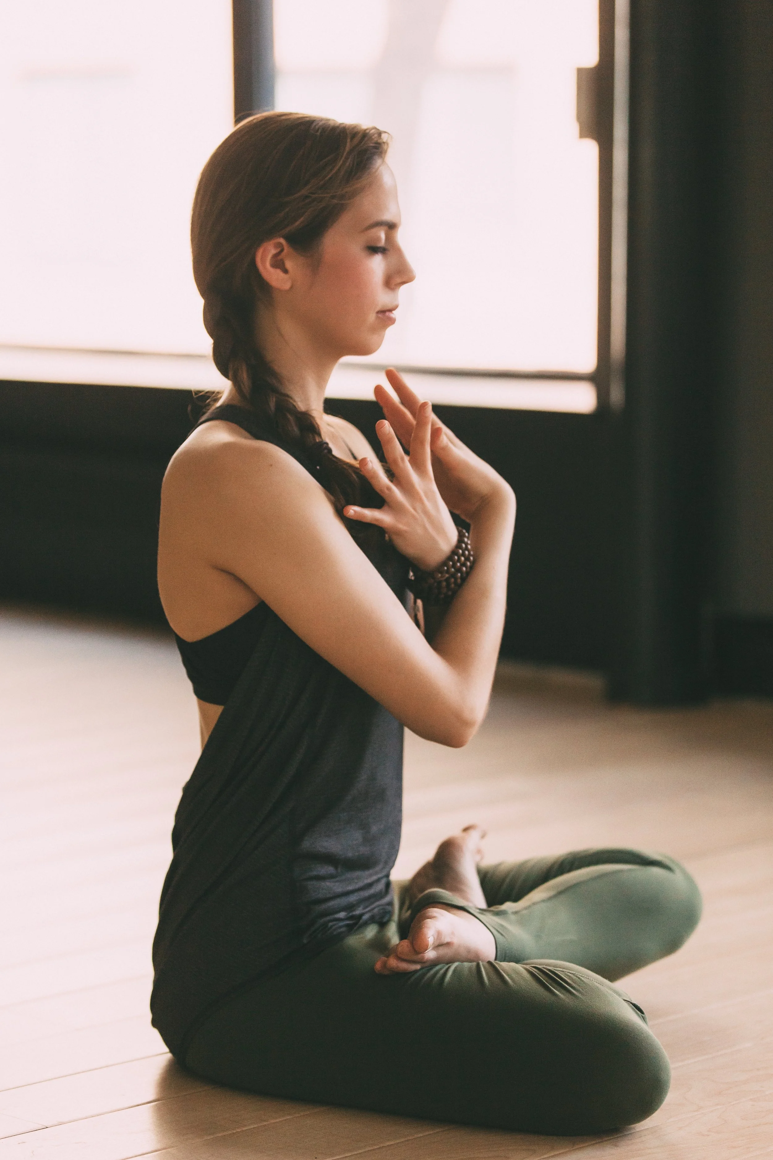 A woman practicing yoga or meditation in a seated cross-legged position with her hands on her chest, eyes closed, wearing a black sports bra and black pants, inside a bright room with a wooden floor and a large window behind her.