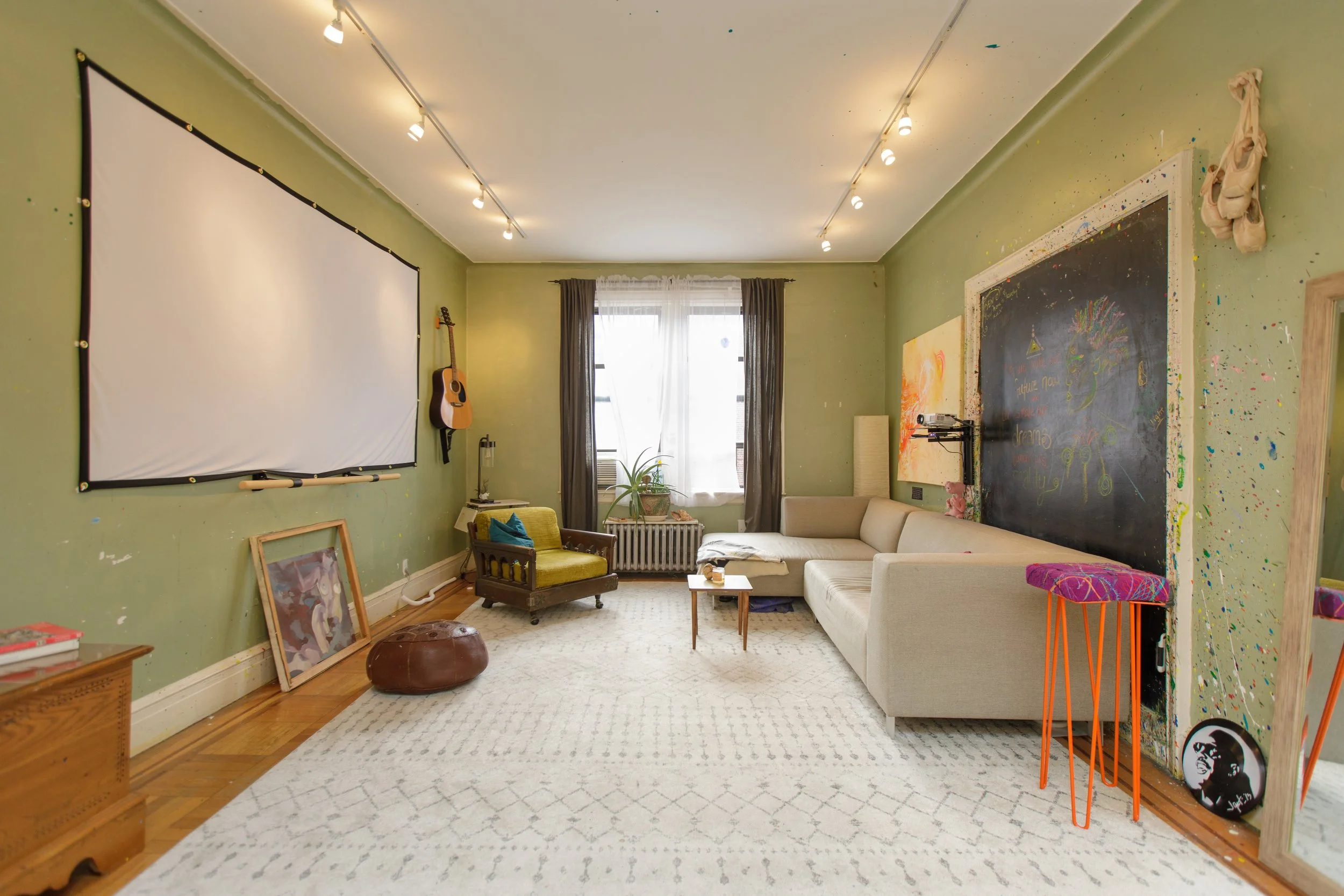 A cozy living room with green painted walls, a large window with dark curtains, and various furniture including a white sectional sofa, a yellow armchair, and a small wooden coffee table. The walls have decorations such as a large chalkboard, paintin