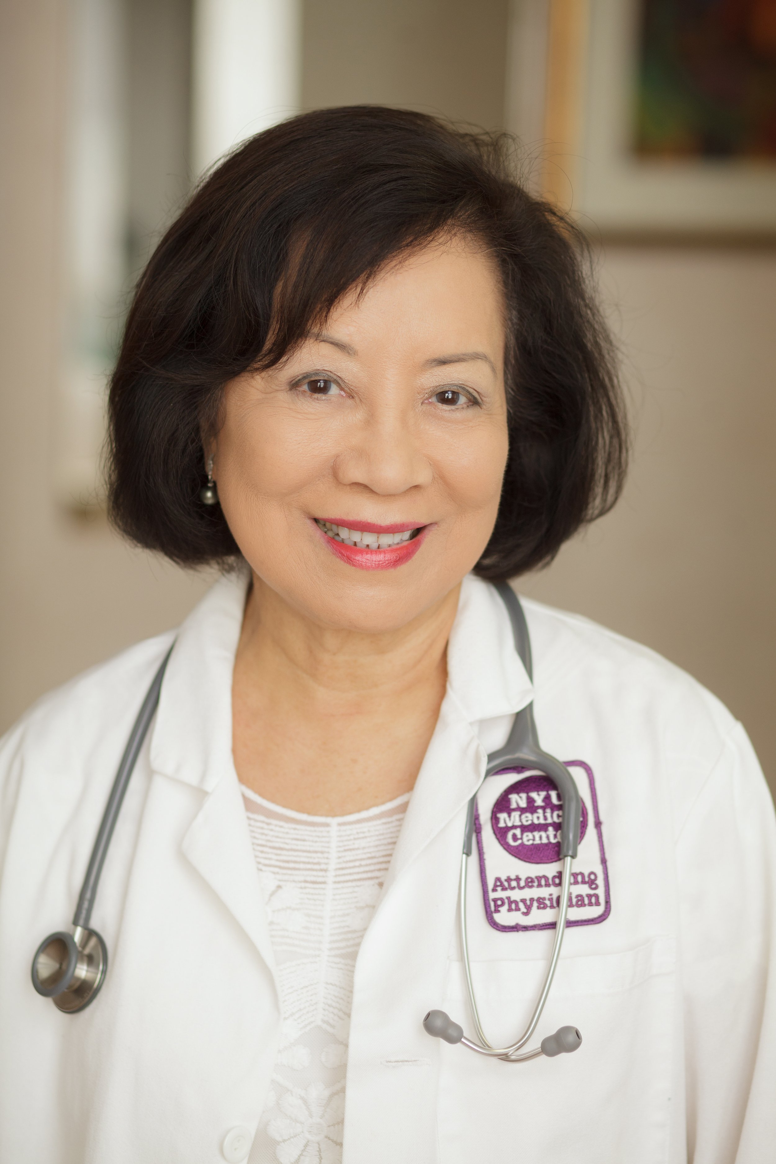 An Asian female doctor smiling, wearing a white lab coat with a stethoscope around her neck, standing indoors with a blurred background.