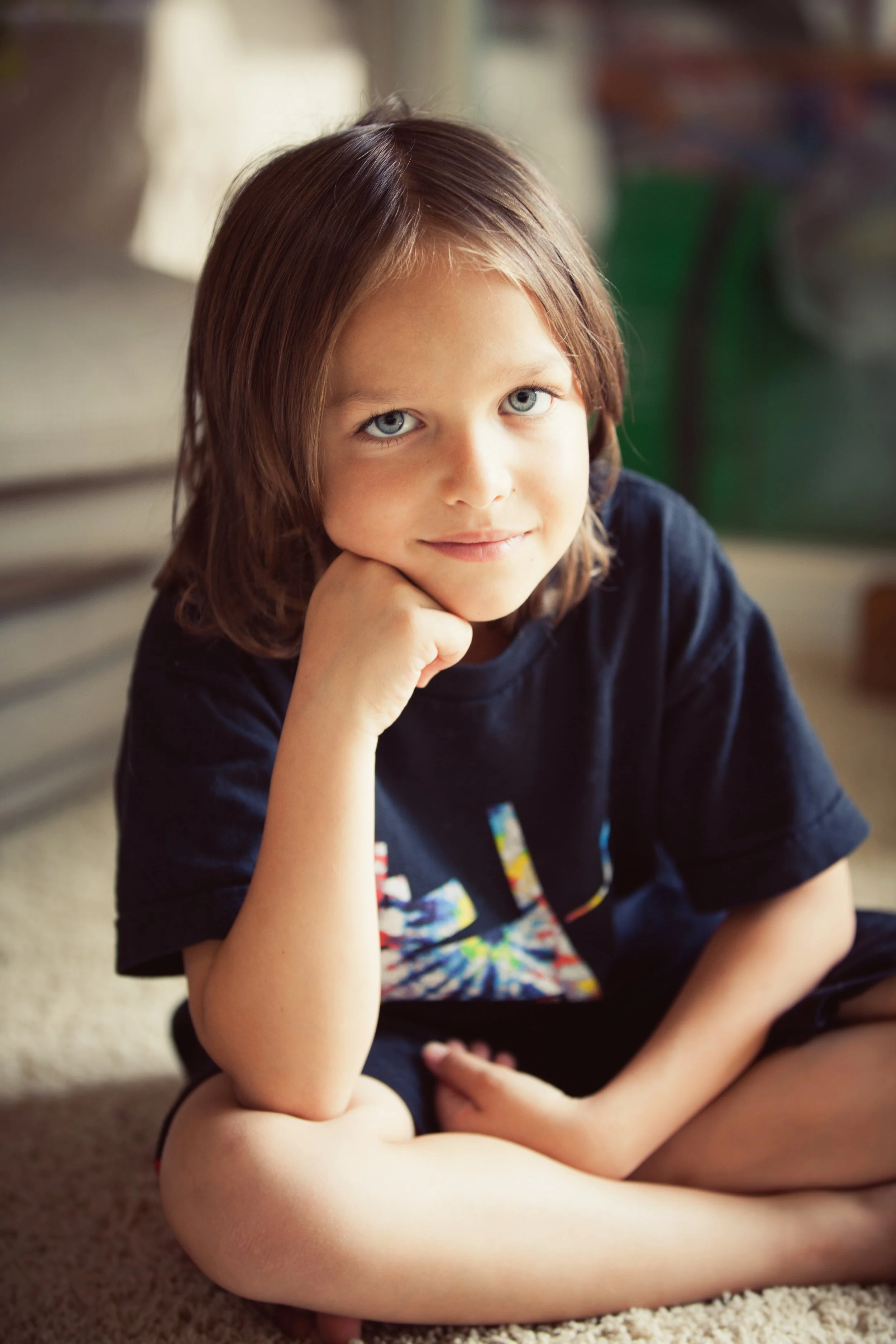 A young boy with long brown hair and blue eyes, sitting on the floor with his hand resting on his chin, smiling softly at the camera.