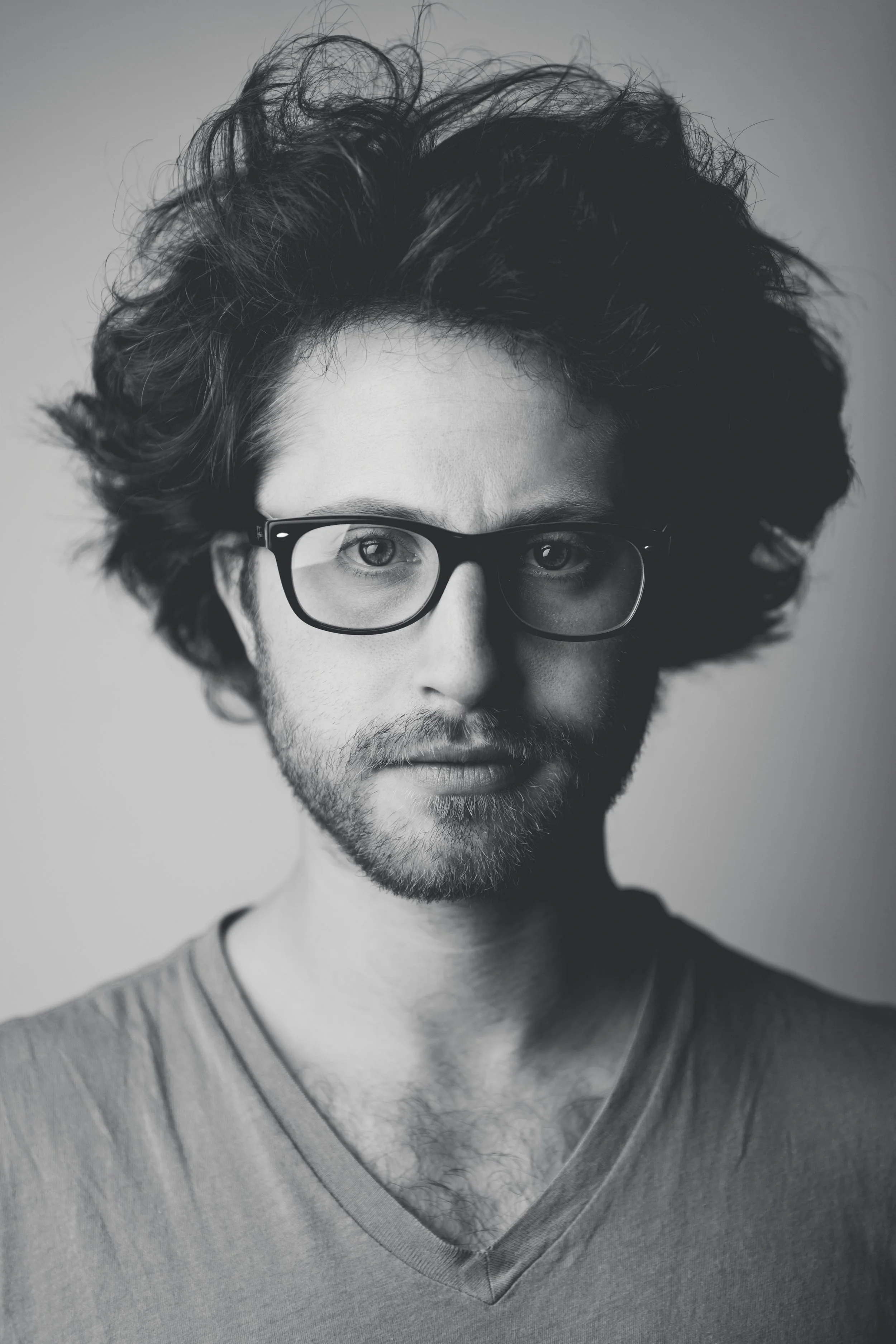Black and white portrait of a young man with curly hair and glasses, wearing a V-neck shirt.