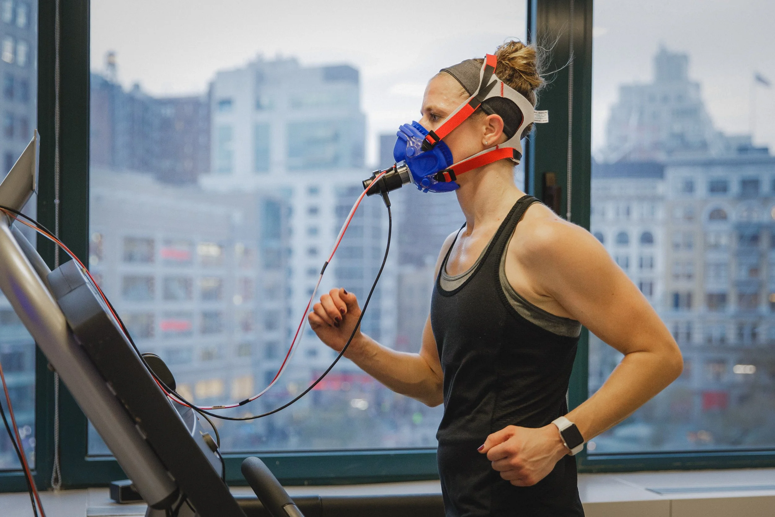 A woman running on a treadmill inside a high-rise building with large windows showing city buildings outside. She is wearing a black tank top, a gray headband, and a respiratory mask connected to exercise sensors.