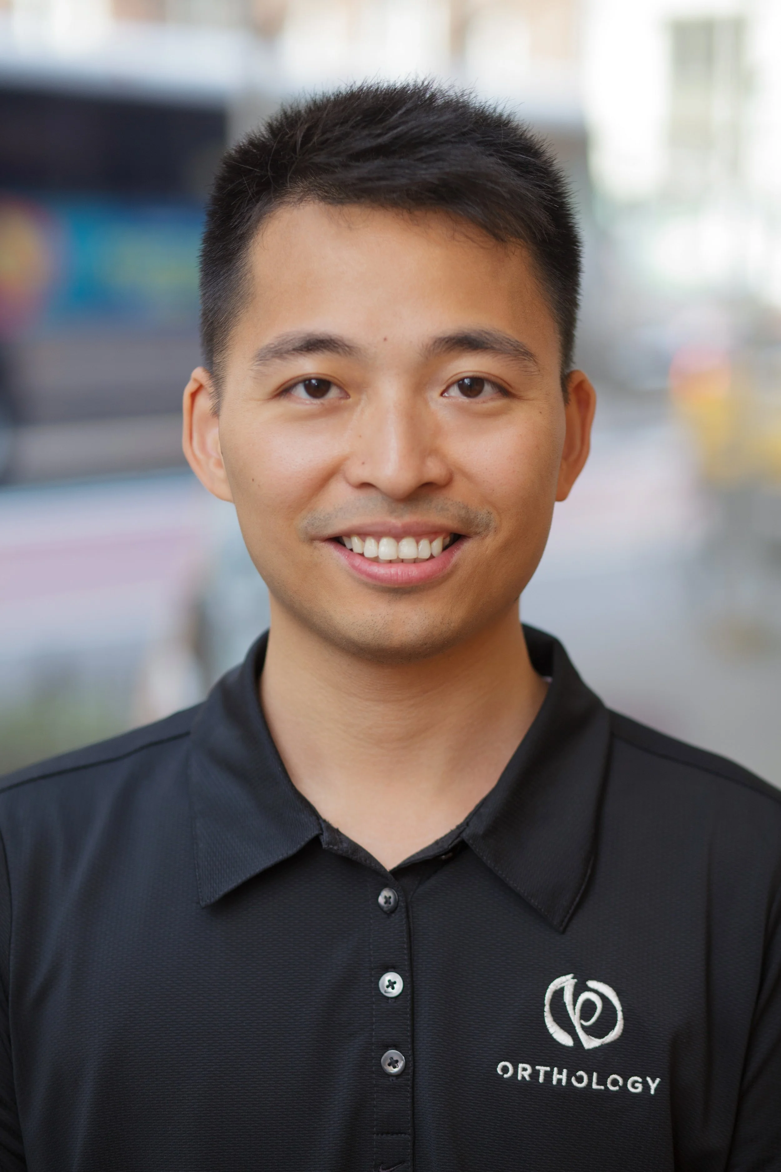 A young man with short black hair smiling, wearing a black orthodontics shirt with a logo that says Orthology.
