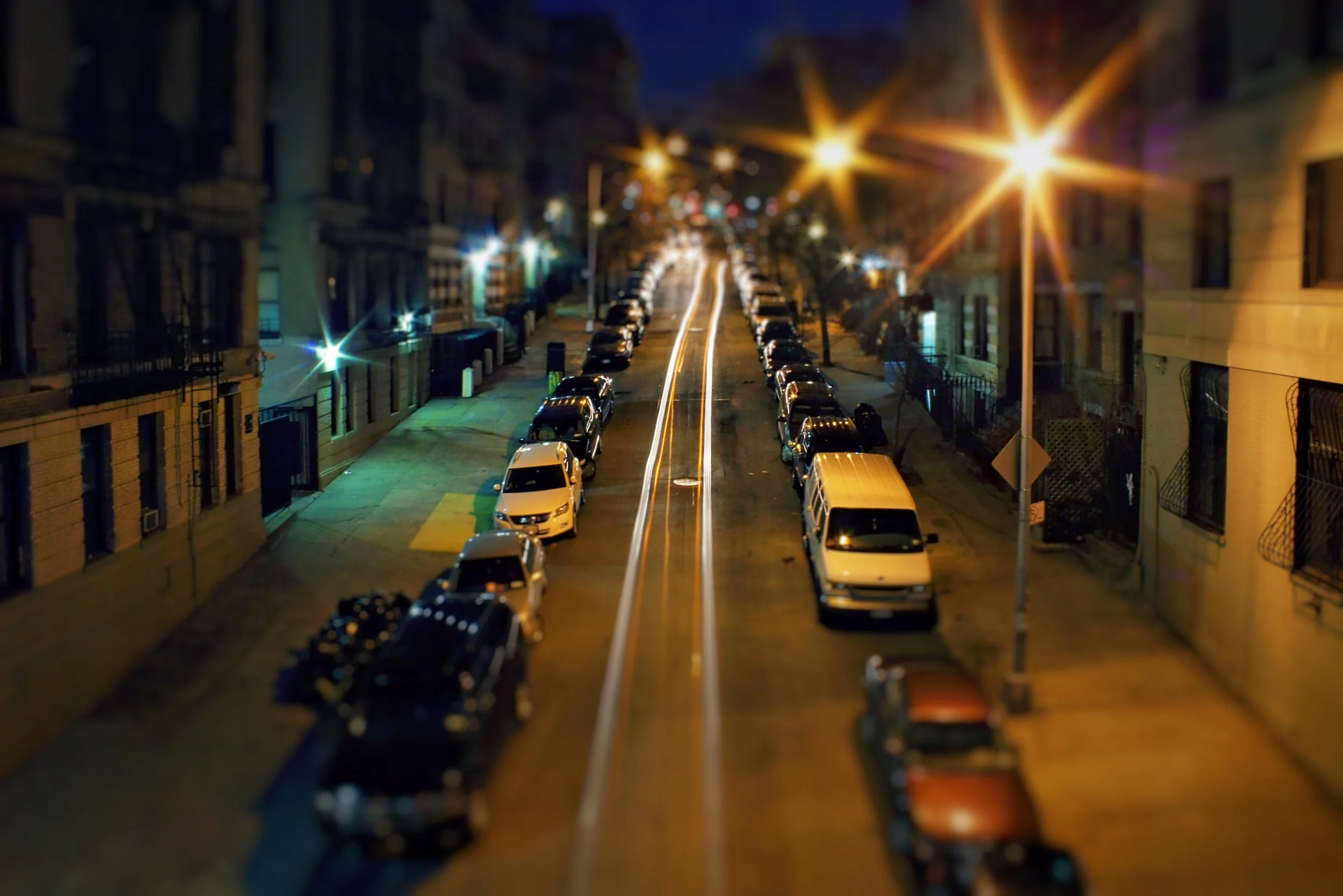 A dimly lit city street at night with parked cars lining both sides. Streetlights cast a warm glow, and the long exposure captures streaks of car light trails down the center of the street.