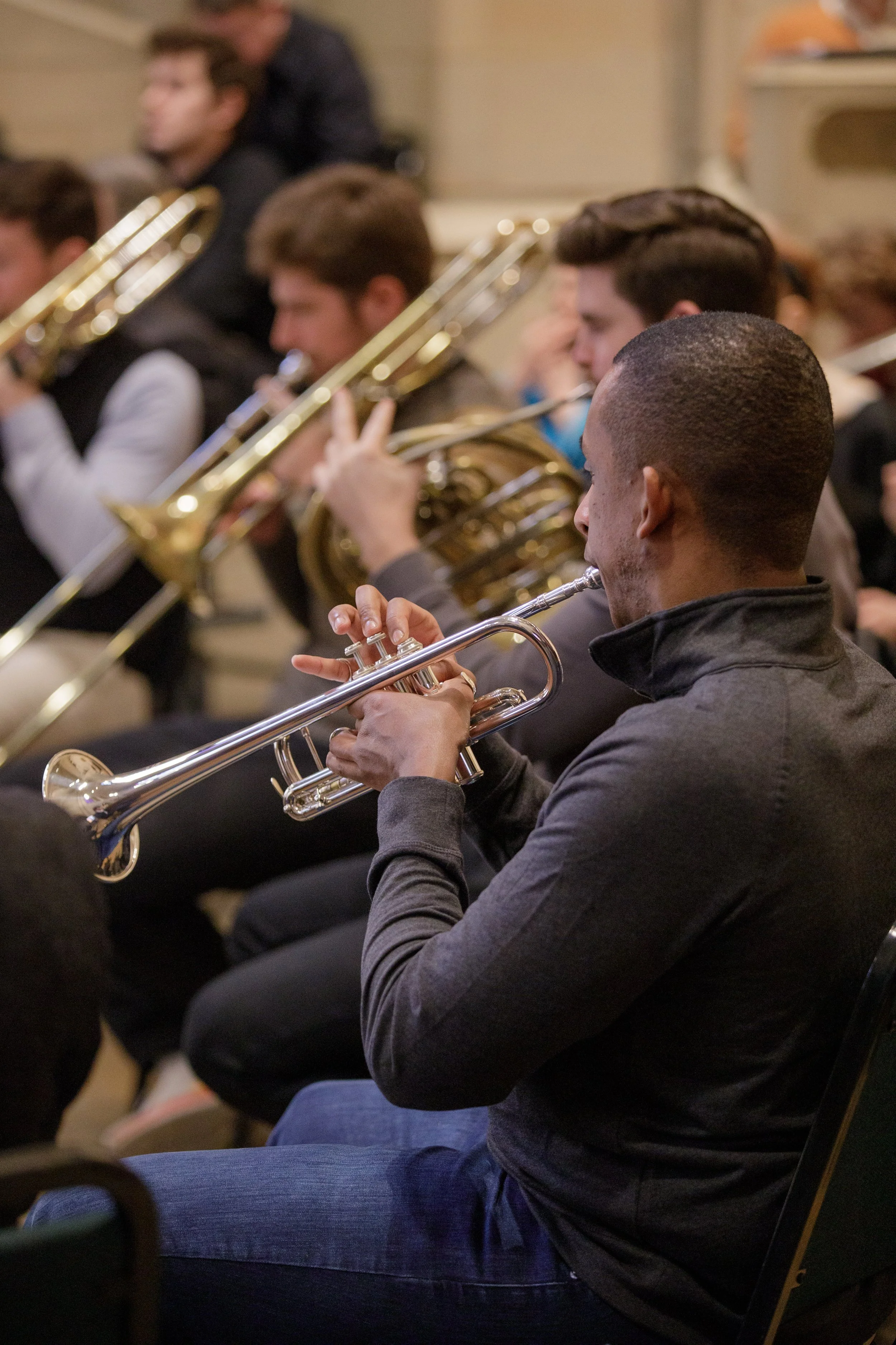 Young man playing the trumpet in an orchestra or band, seated among other musicians, in an indoor setting.