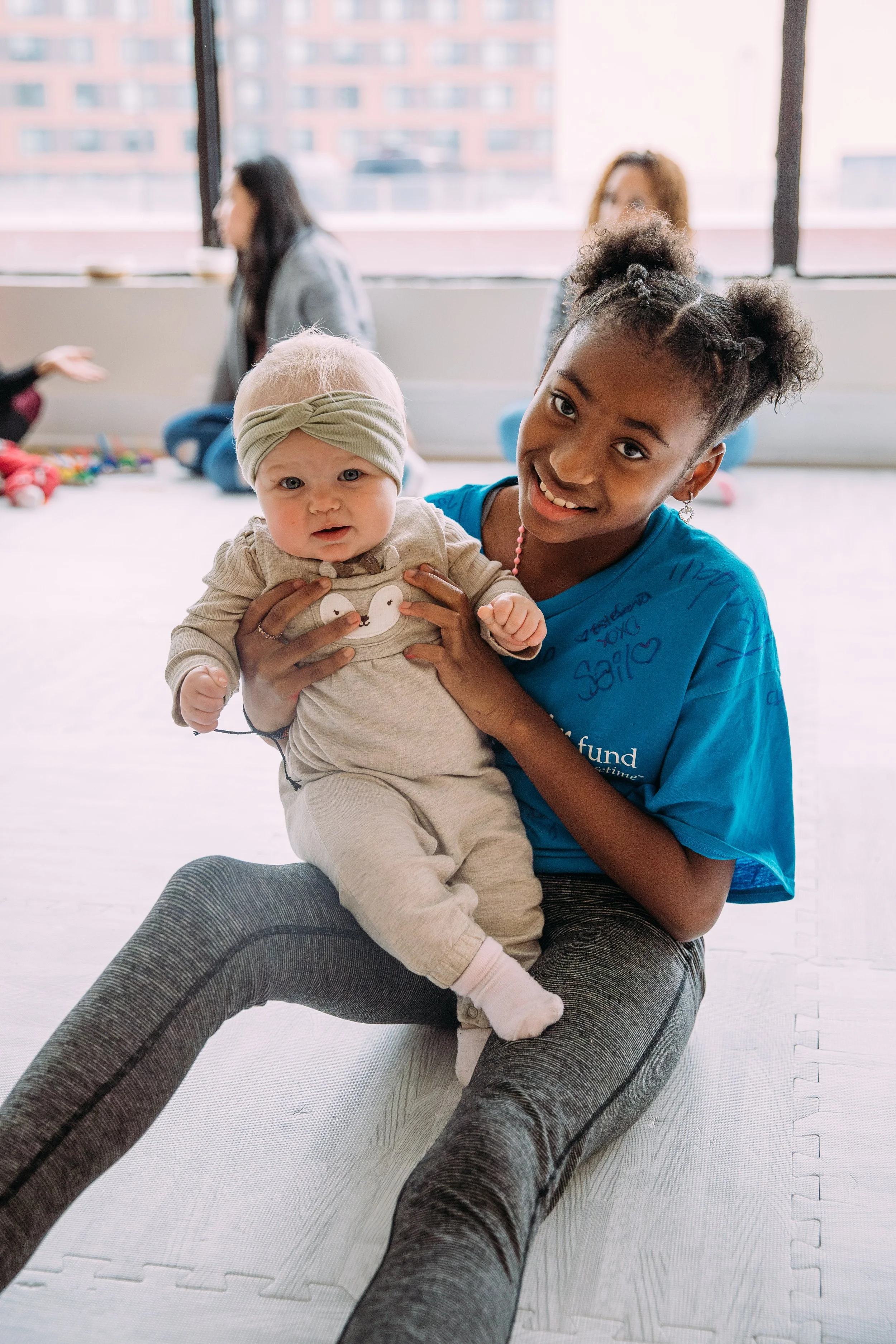 A young girl with curly hair and a school uniform holding a baby, sitting on the floor in a bright room with large windows, with other women blurred in the background.