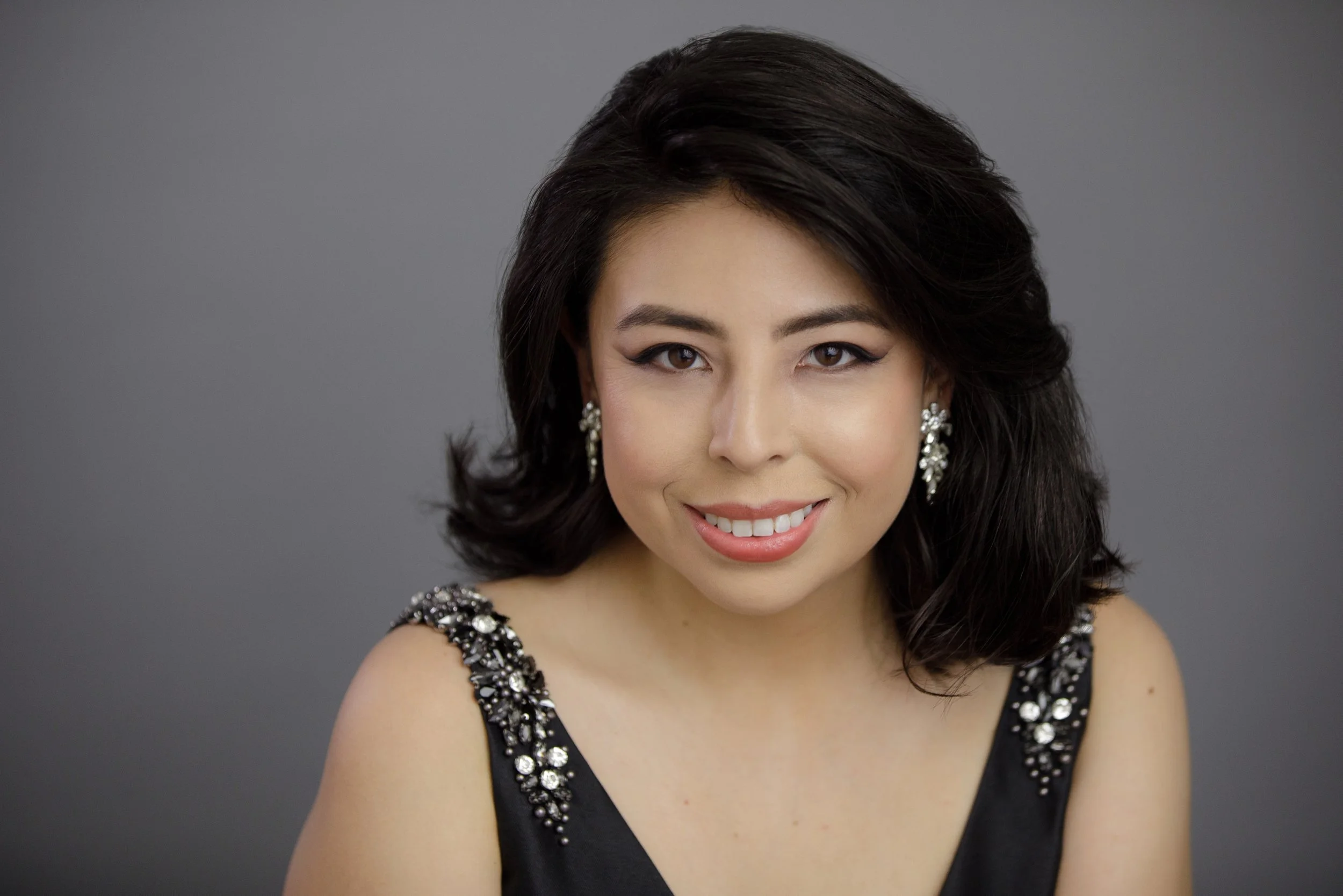 A woman with dark hair styled in loose waves, wearing pearl and rhinestone earrings, and a black dress with jeweled embellishments on the shoulders, smiling against a gray background.