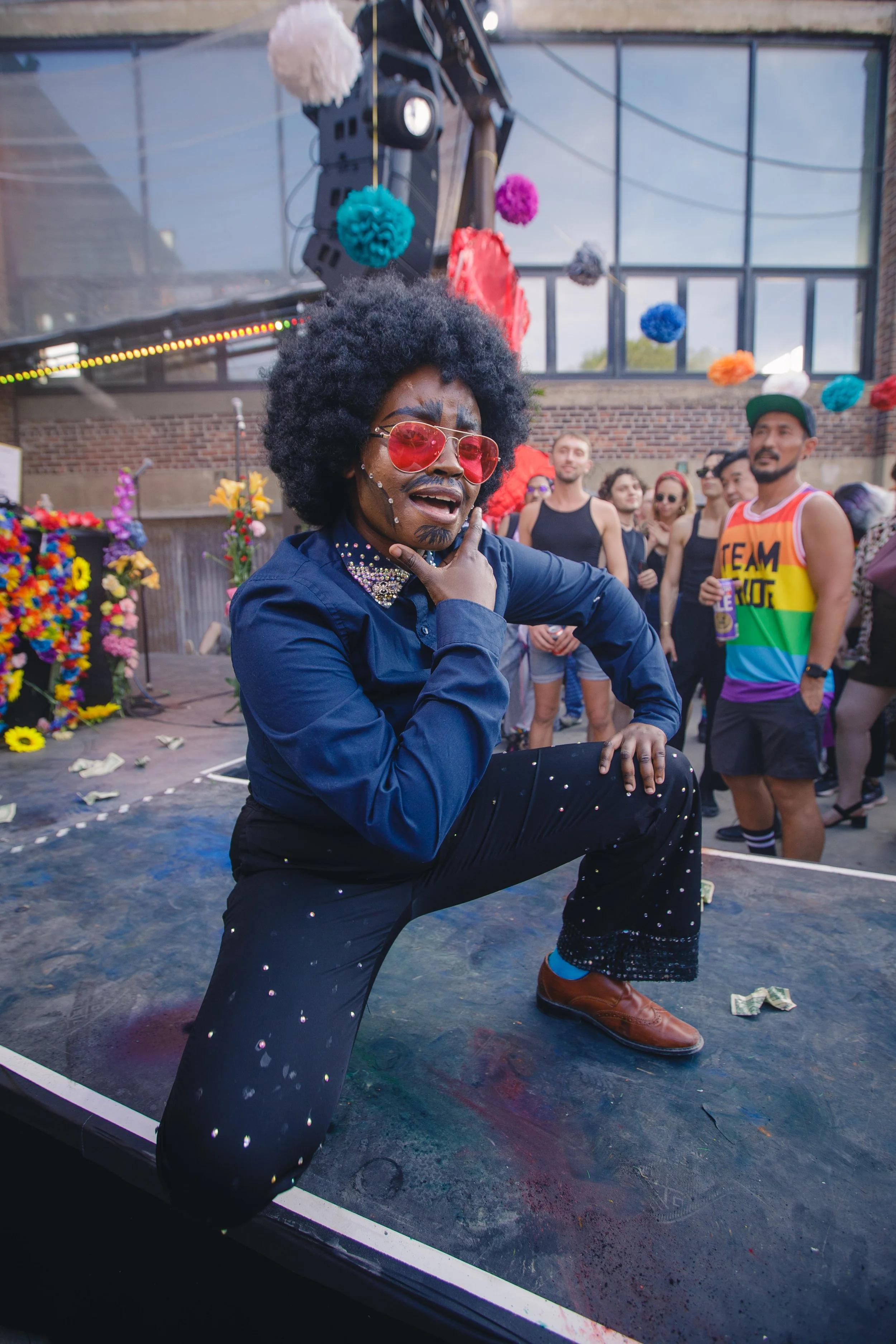 A person dressed in a dark blue shirt and black pants with shiny decorations, wearing pink sunglasses, is posing for the camera at a lively outdoor event with colorful paper decorations, flowers, and a crowd of people in the background.