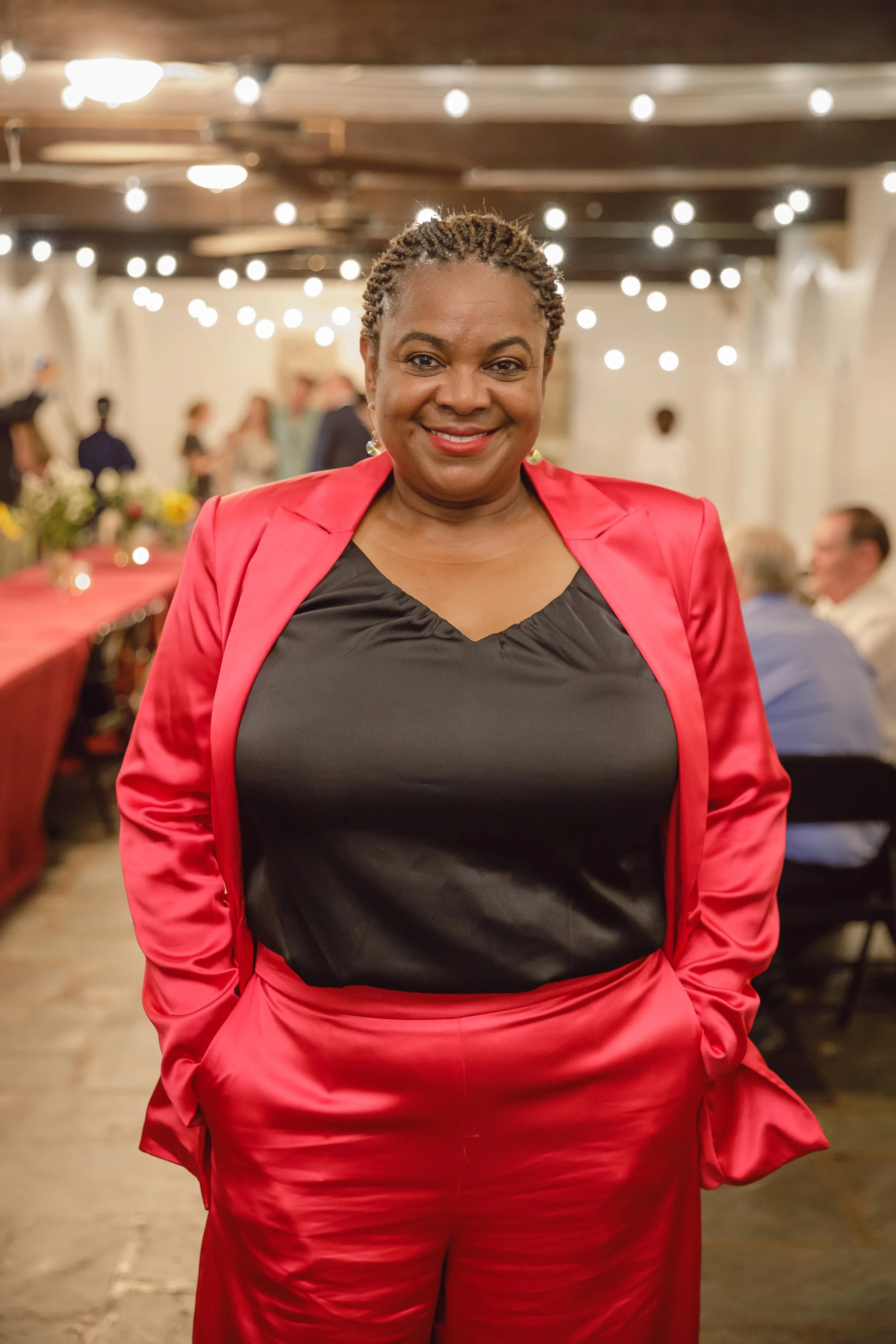 A woman smiling at a social gathering, wearing a red satin suit with a black top underneath, with blurred background of other guests, string lights, and a decorated table.