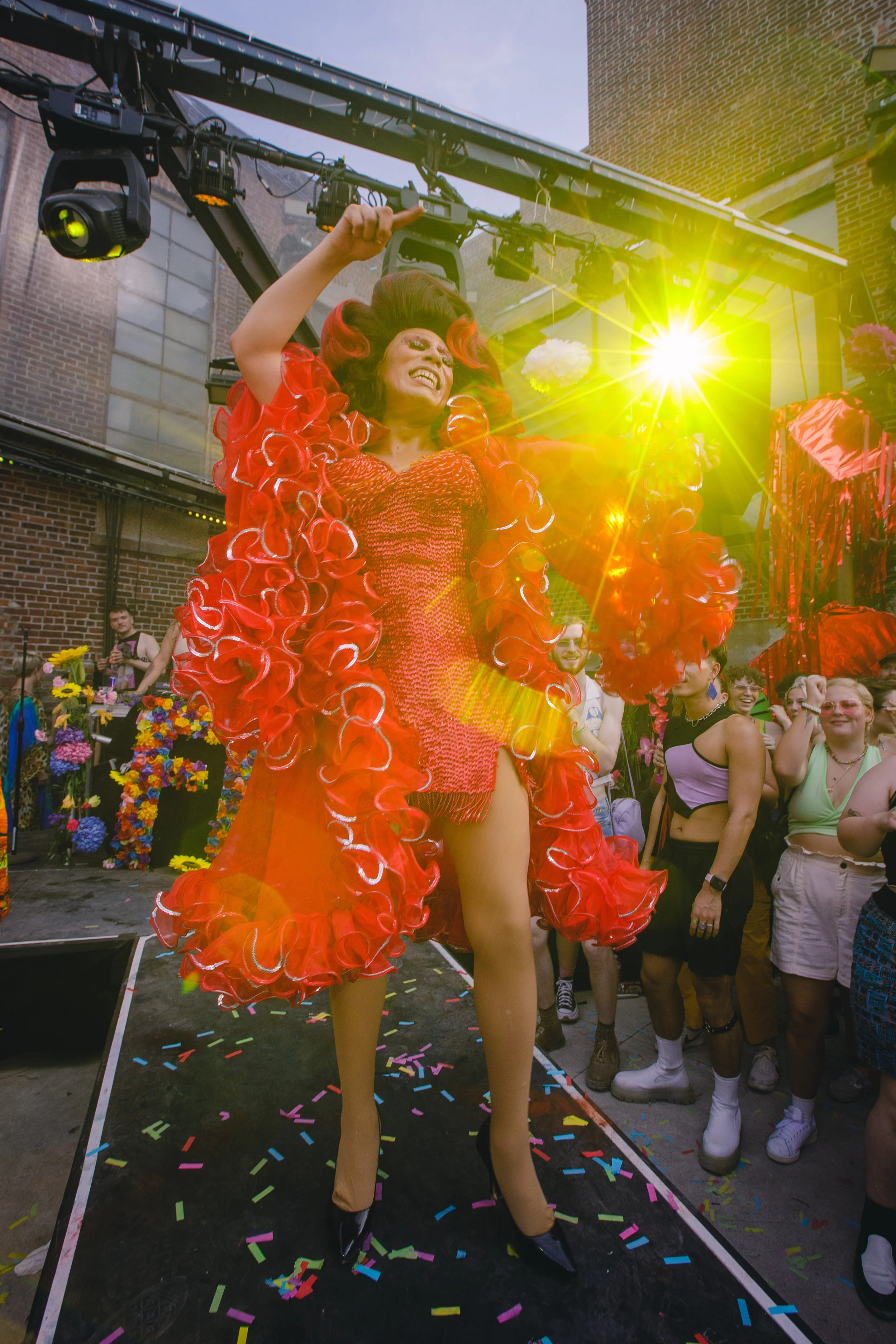 Performer in a red, ruffled outfit dancing on a stage during an outdoor event, surrounded by a crowd of cheering spectators with brick buildings in the background.