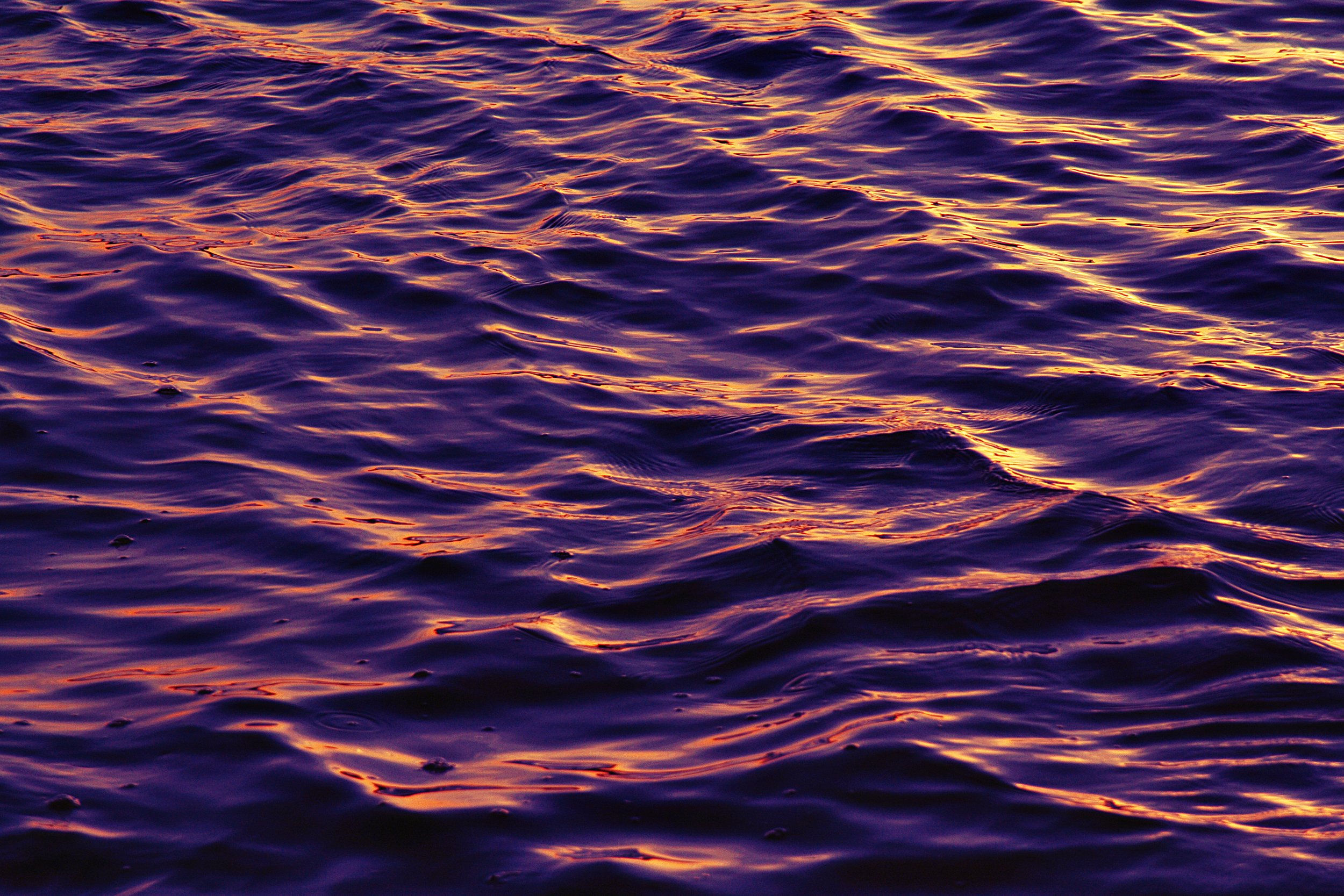 Close-up of water surface with ripples reflecting a vibrant sunset or sunrise in shades of purple, orange, and gold.