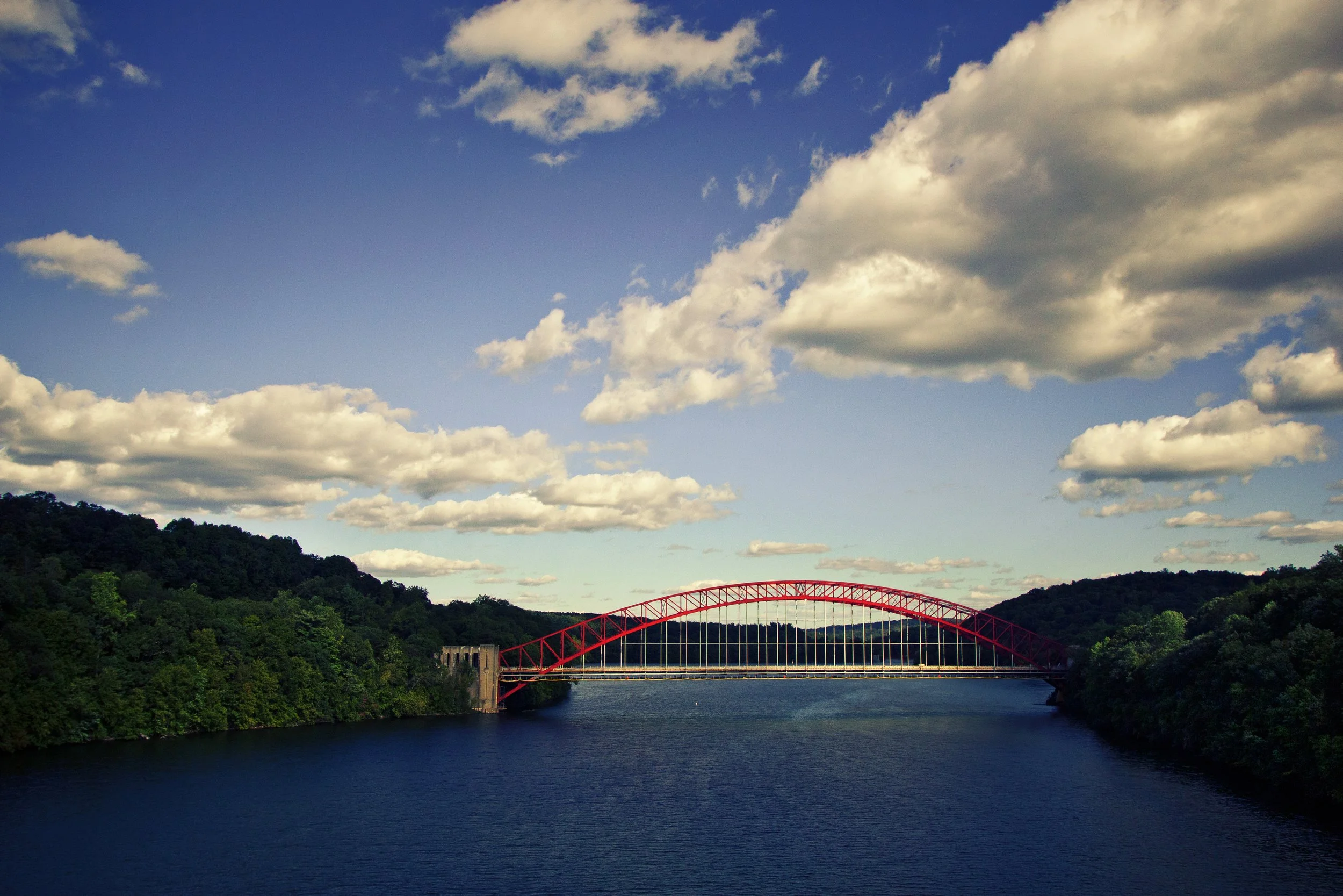 A red arched bridge spanning over a body of water, surrounded by green hills with scattered trees, under a blue sky with white clouds.