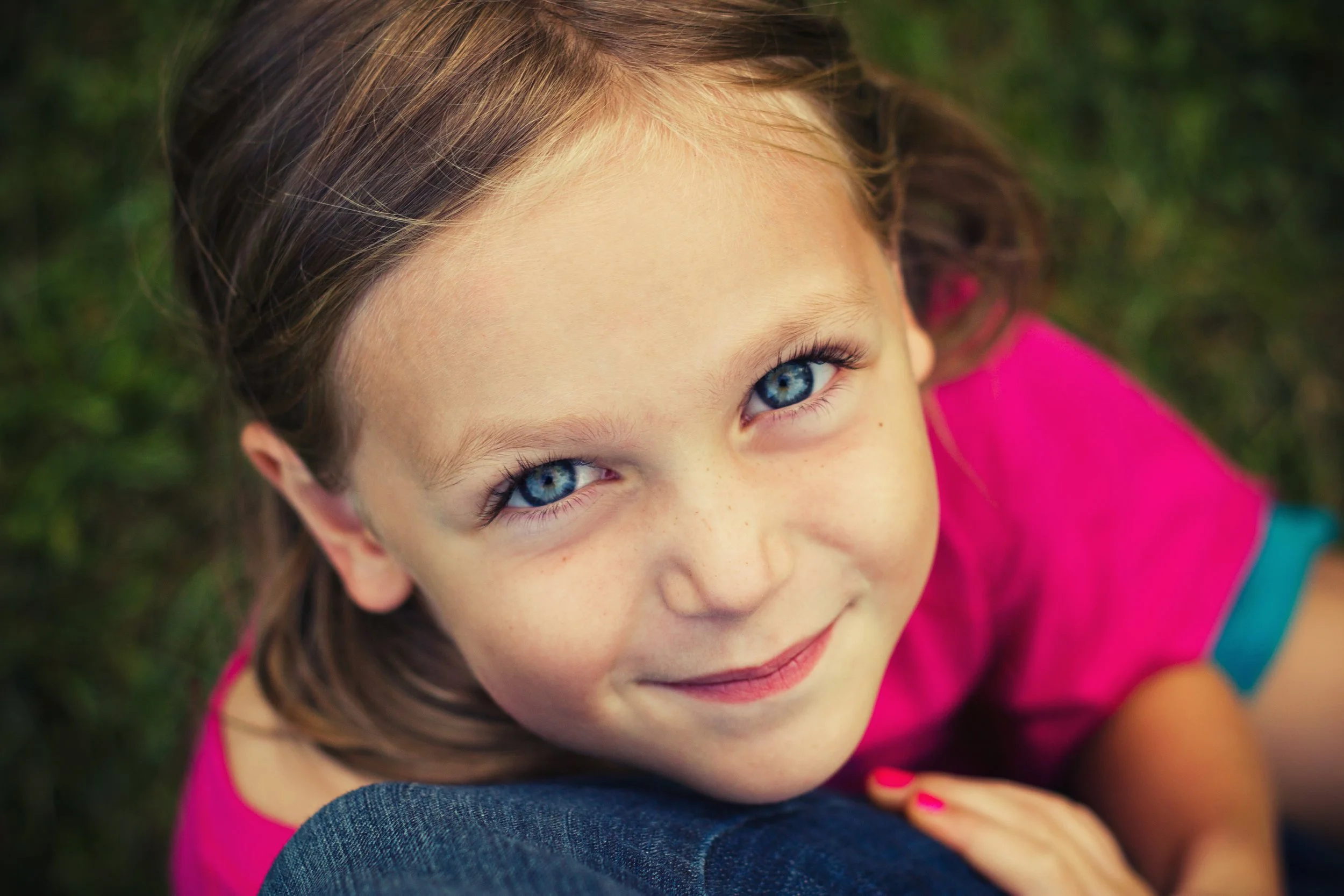 Close-up of a young girl with blue eyes and light brown hair, gazing upward, outdoors with blurred green background, wearing a pink top.