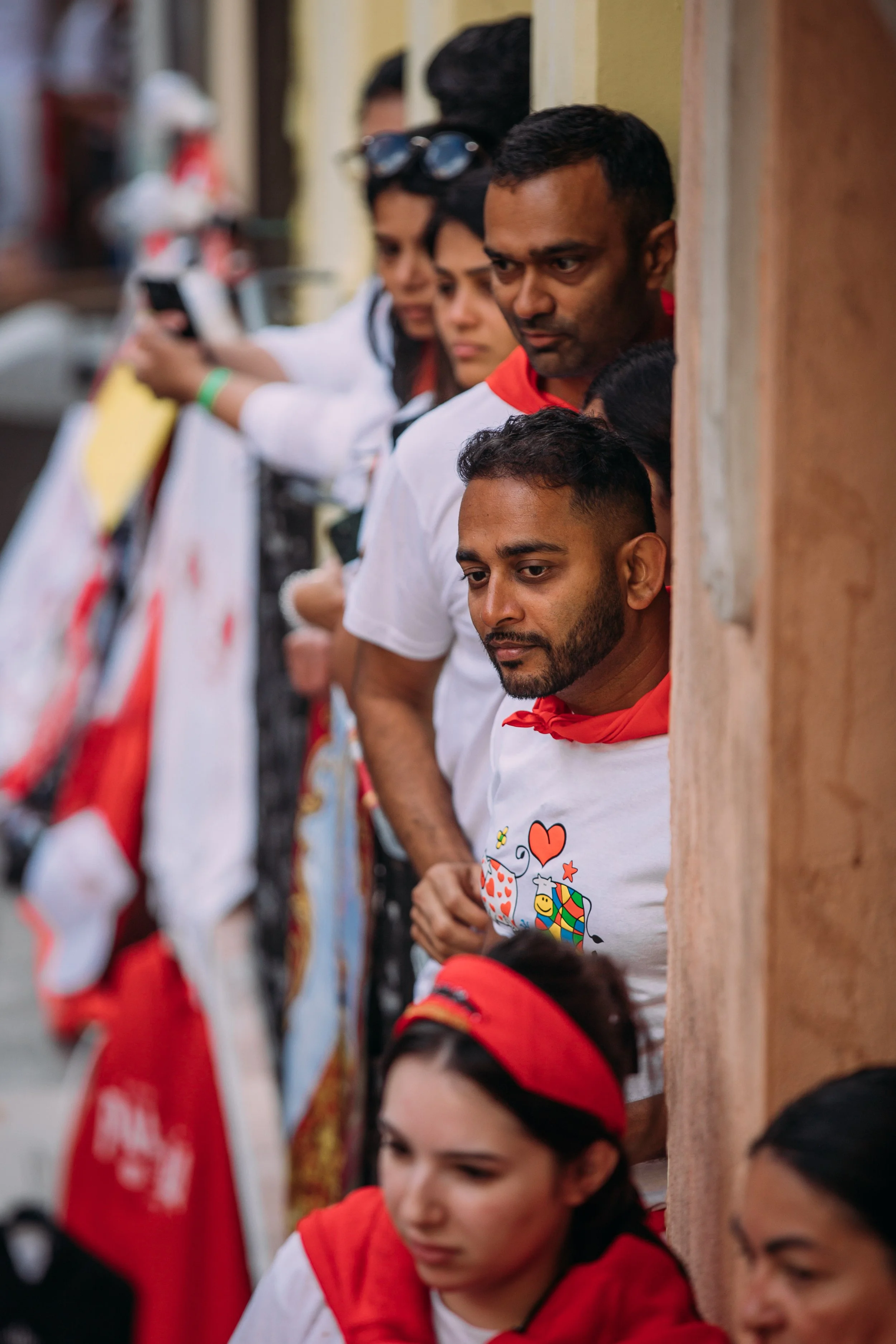 People are standing in a line, with their heads peeking through a wall opening, some wearing red shirts and scarves, in an outdoor setting.