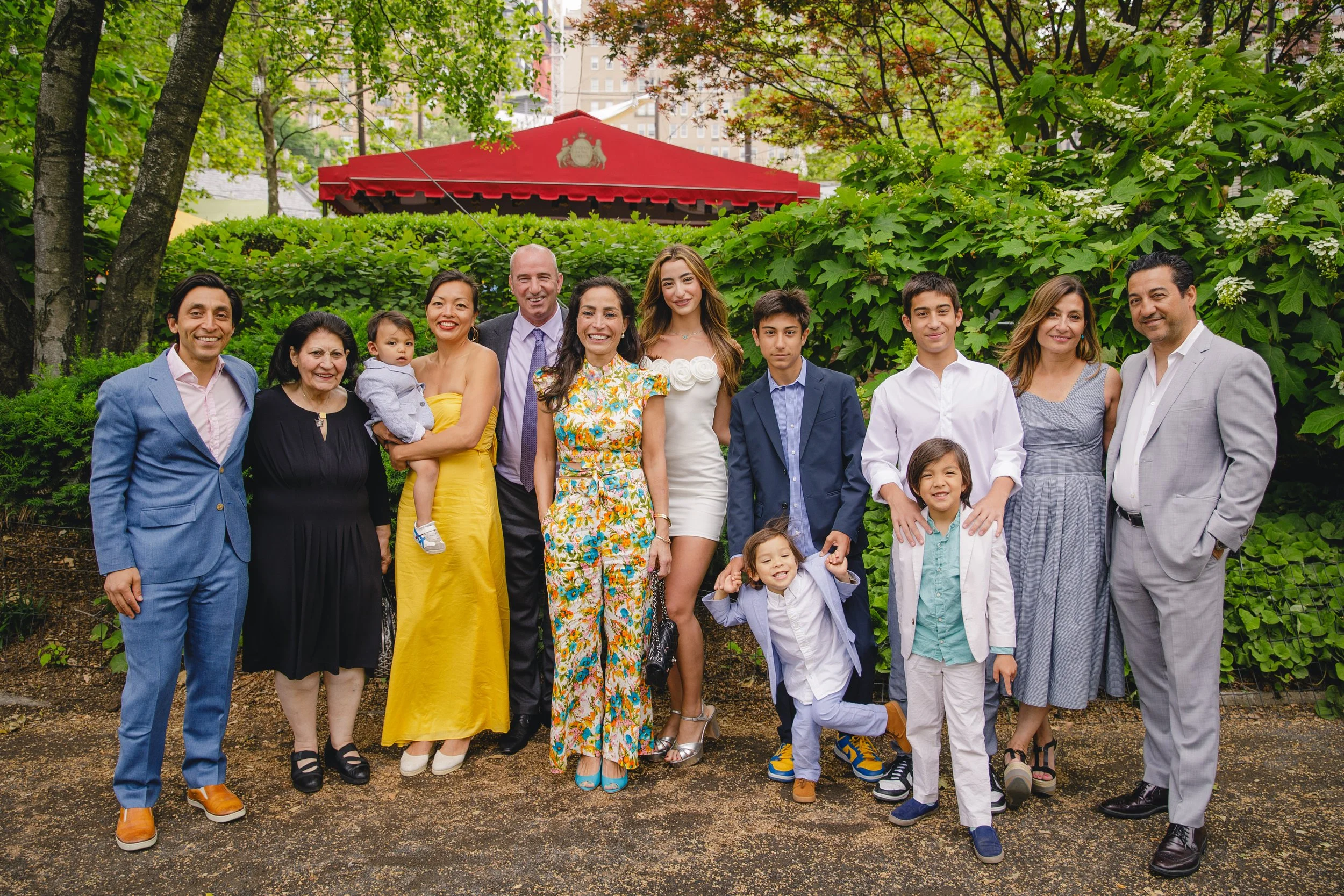 A large group of diverse people, including adults and children, standing outdoors in a park with green trees and bushes, smiling for a photo, with city buildings in the background.