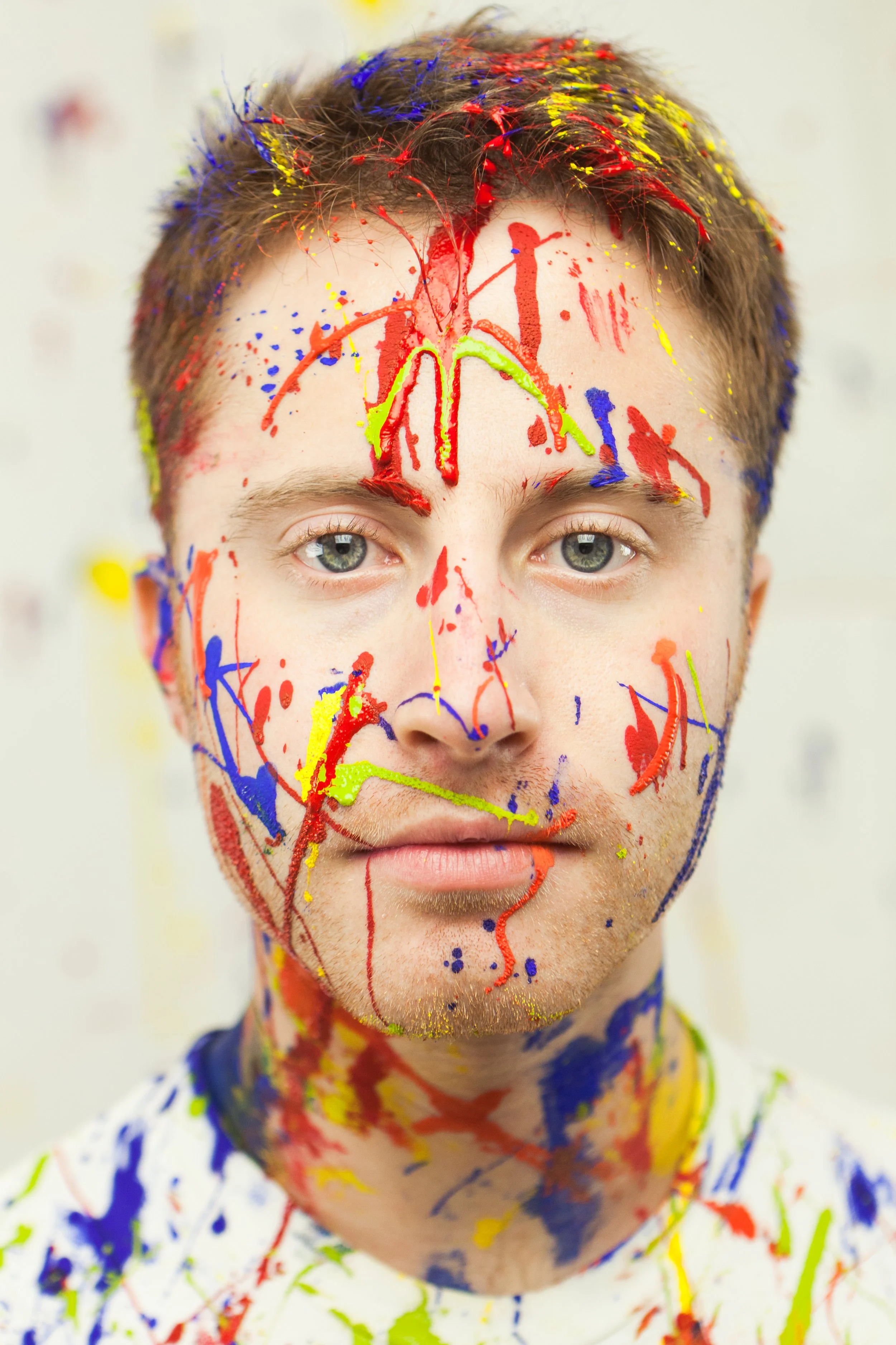 A close-up of a young man with short brown hair and blue eyes, covered in colorful paint splashes and streaks in red, blue, yellow, and green on his face and upper body, with a neutral expression.