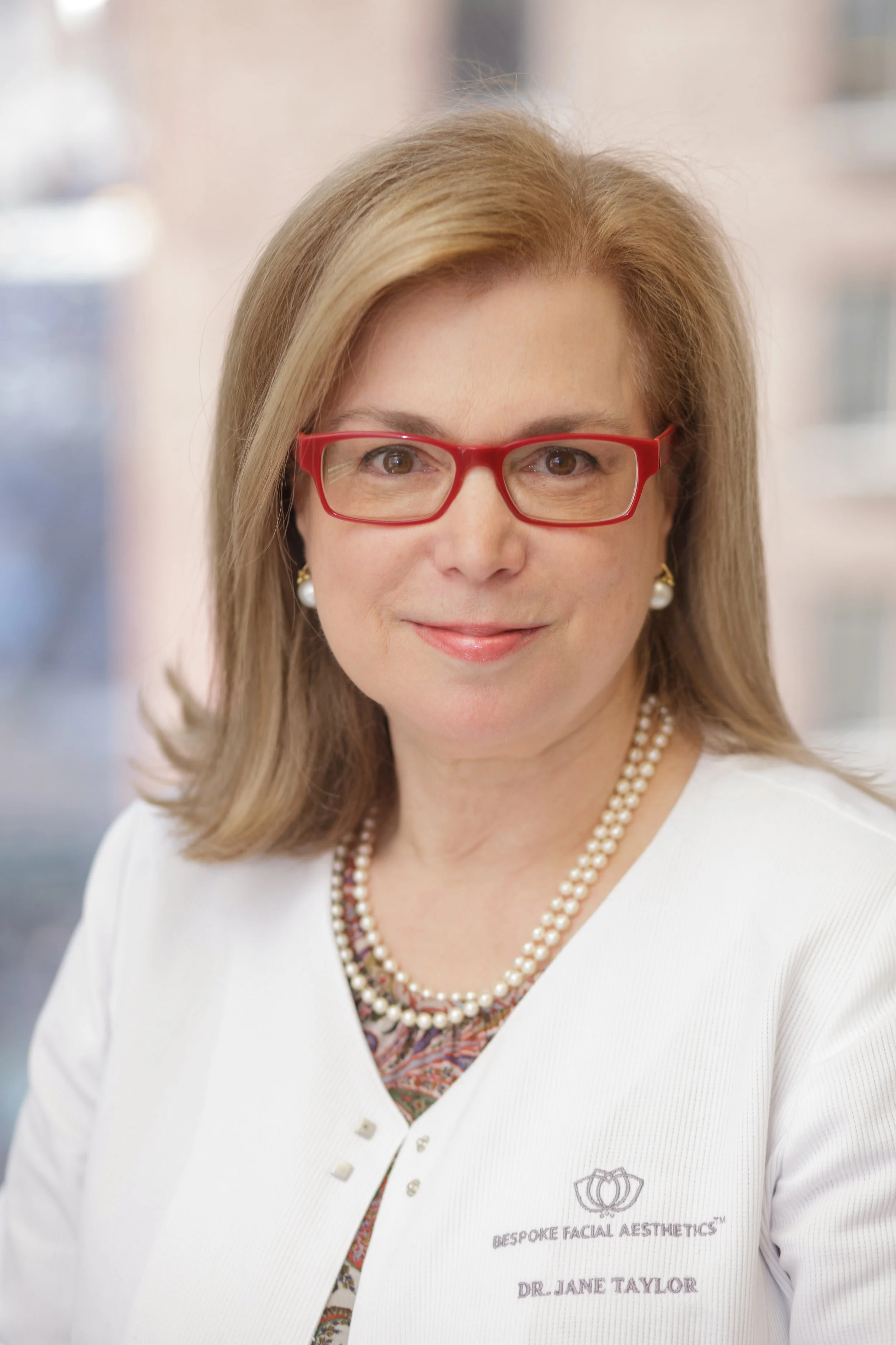 Close-up of a woman wearing red glasses, pearl earrings, and a white coat with a name tag that reads 'Dr. Jane Taylor' and the logo 'Bespoke Facial Aesthetics.' She has shoulder-length blonde hair, and a background of an indoor setting.