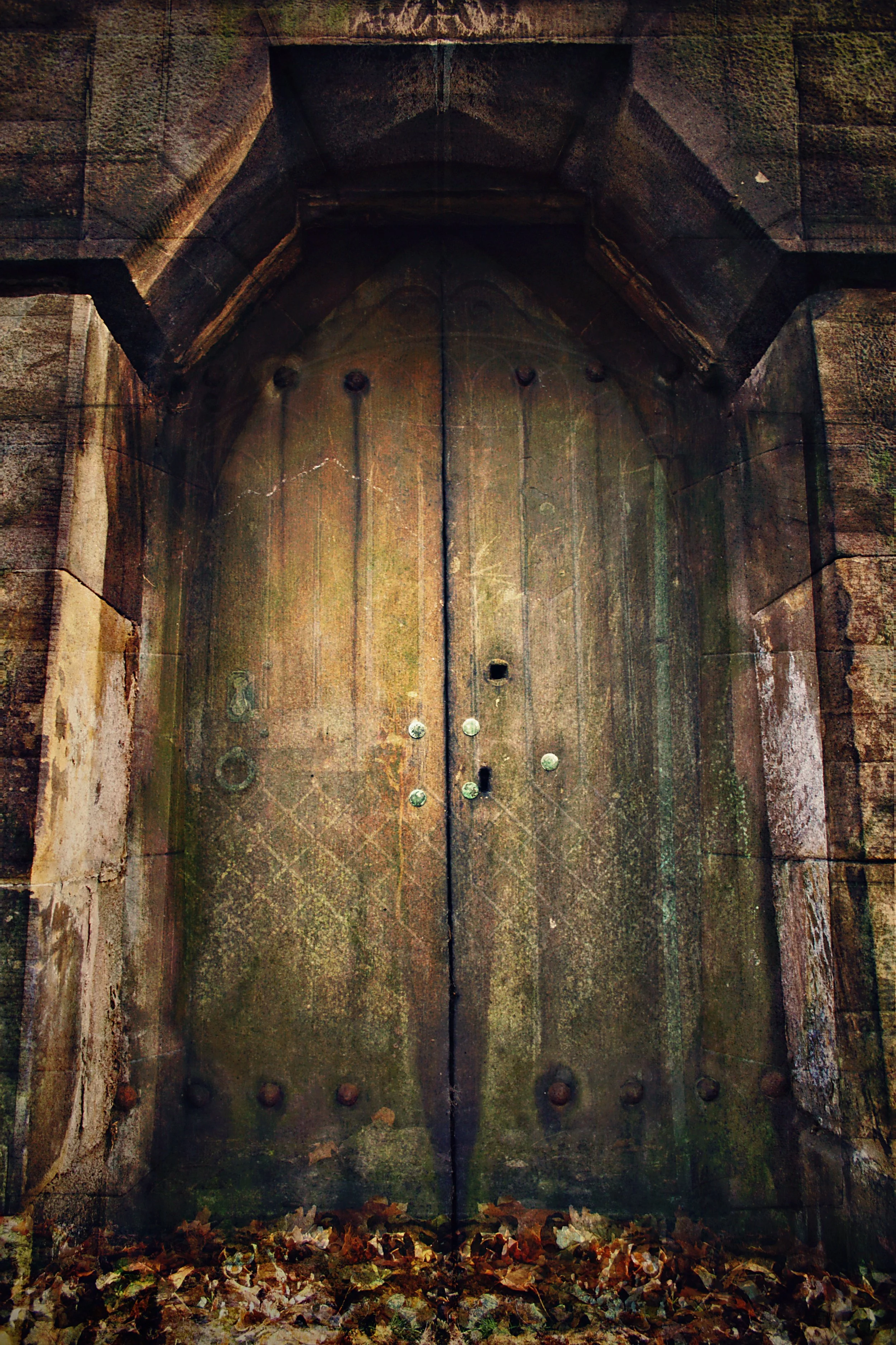 An old, large, heavy wooden door with metal studs, set in a stone wall, with fallen leaves at its base.