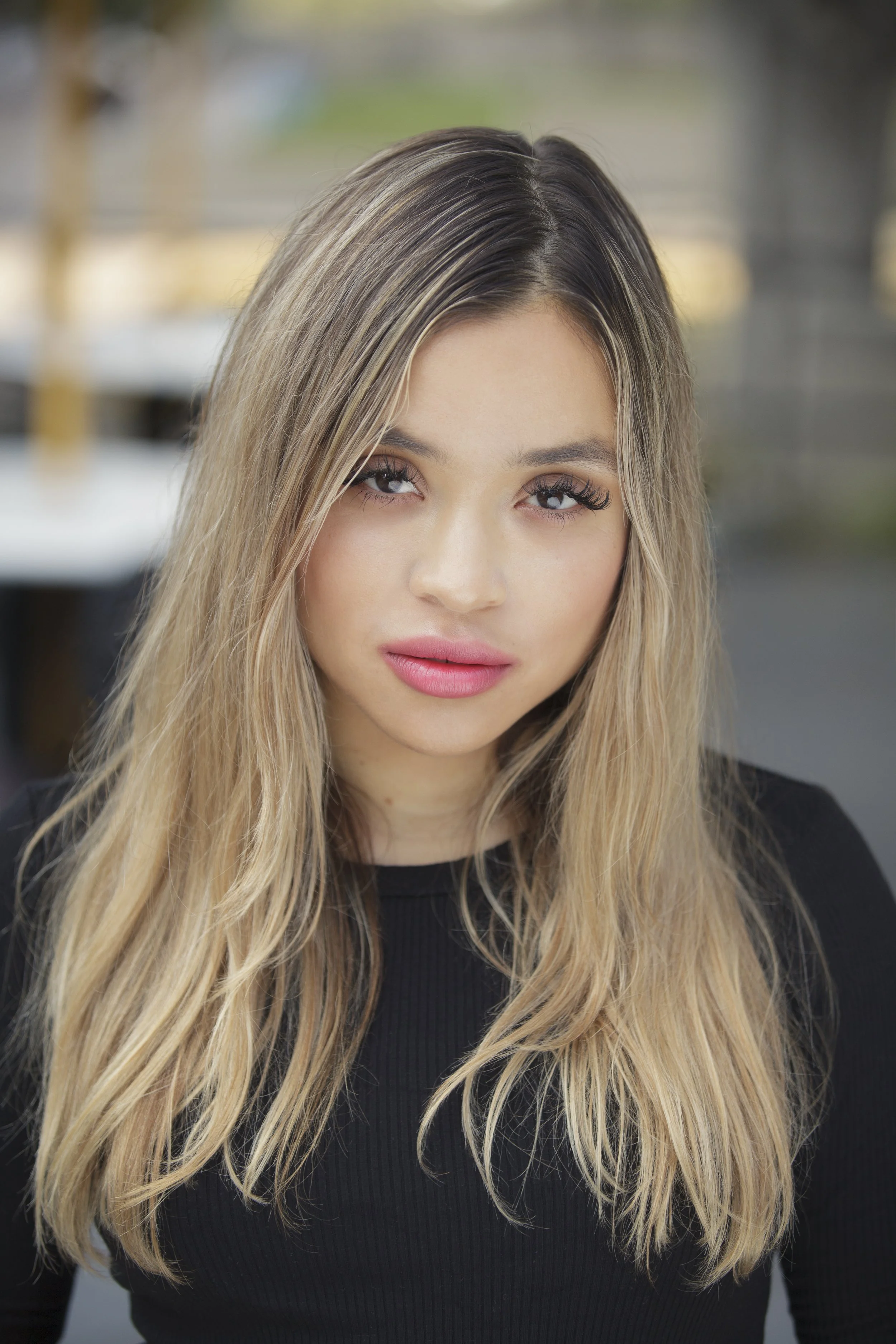 A young woman with long, wavy blonde hair and makeup, wearing a black top, looking at the camera with a blurred outdoor background.