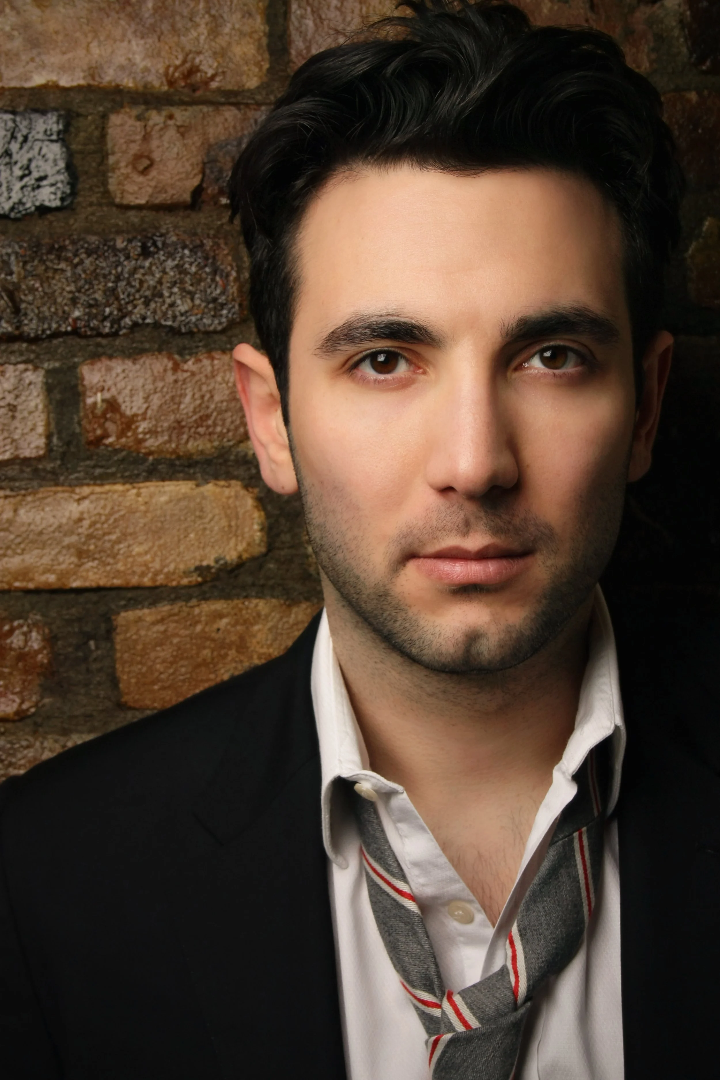 Close-up of a man with dark hair, brown eyes, and light skin, wearing a white shirt, a black blazer, and a gray and red striped tie, standing against a brick wall background.