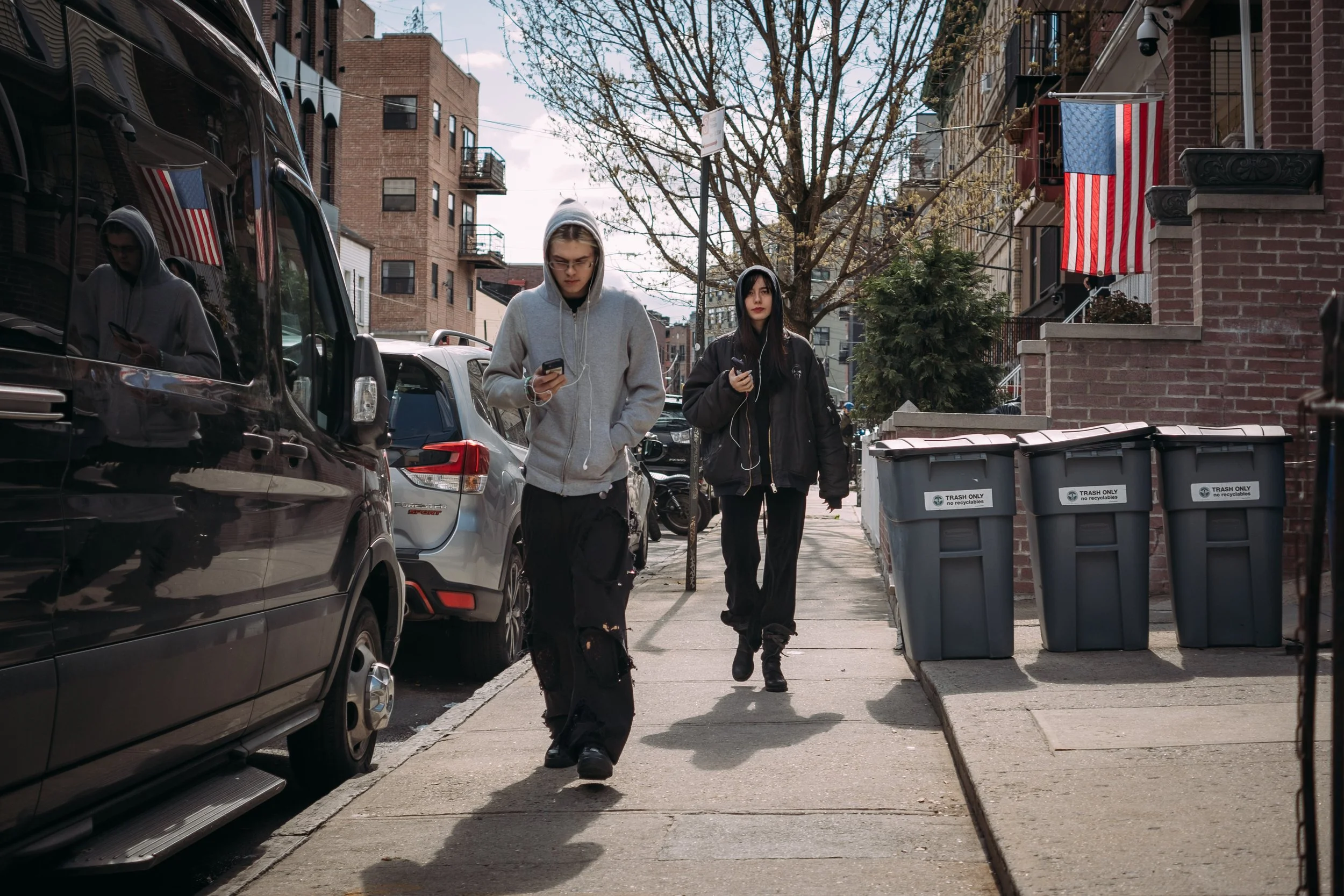 Two young people, a man and a woman, walking on a city street while looking at their phones, with parked cars, residential buildings, and leafless trees in the background.