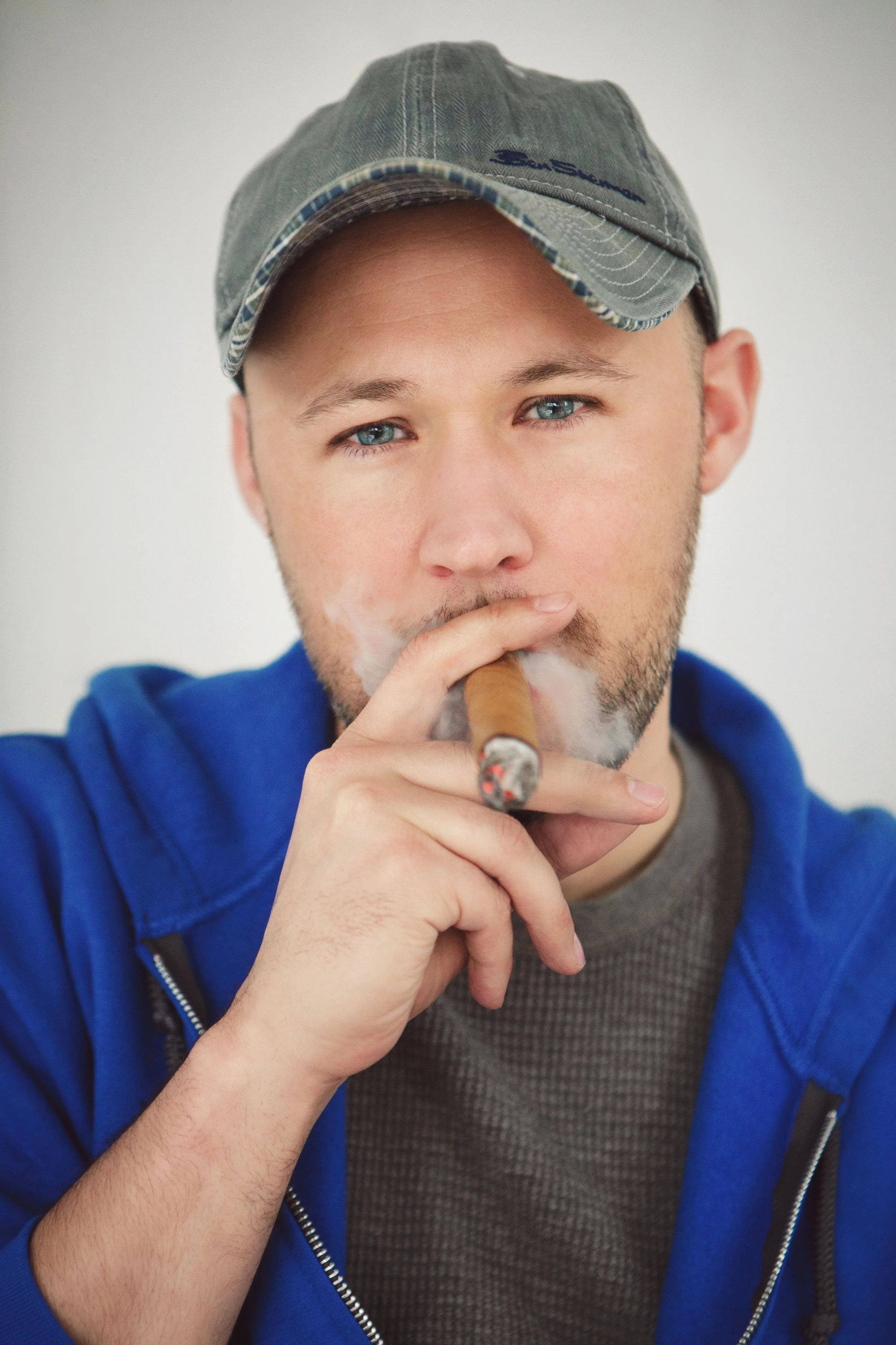 A young man with blue eyes and light facial hair, wearing a gray baseball cap with writing on it, smoking a cigar, and dressed in a blue hoodie and gray shirt, against a plain background.