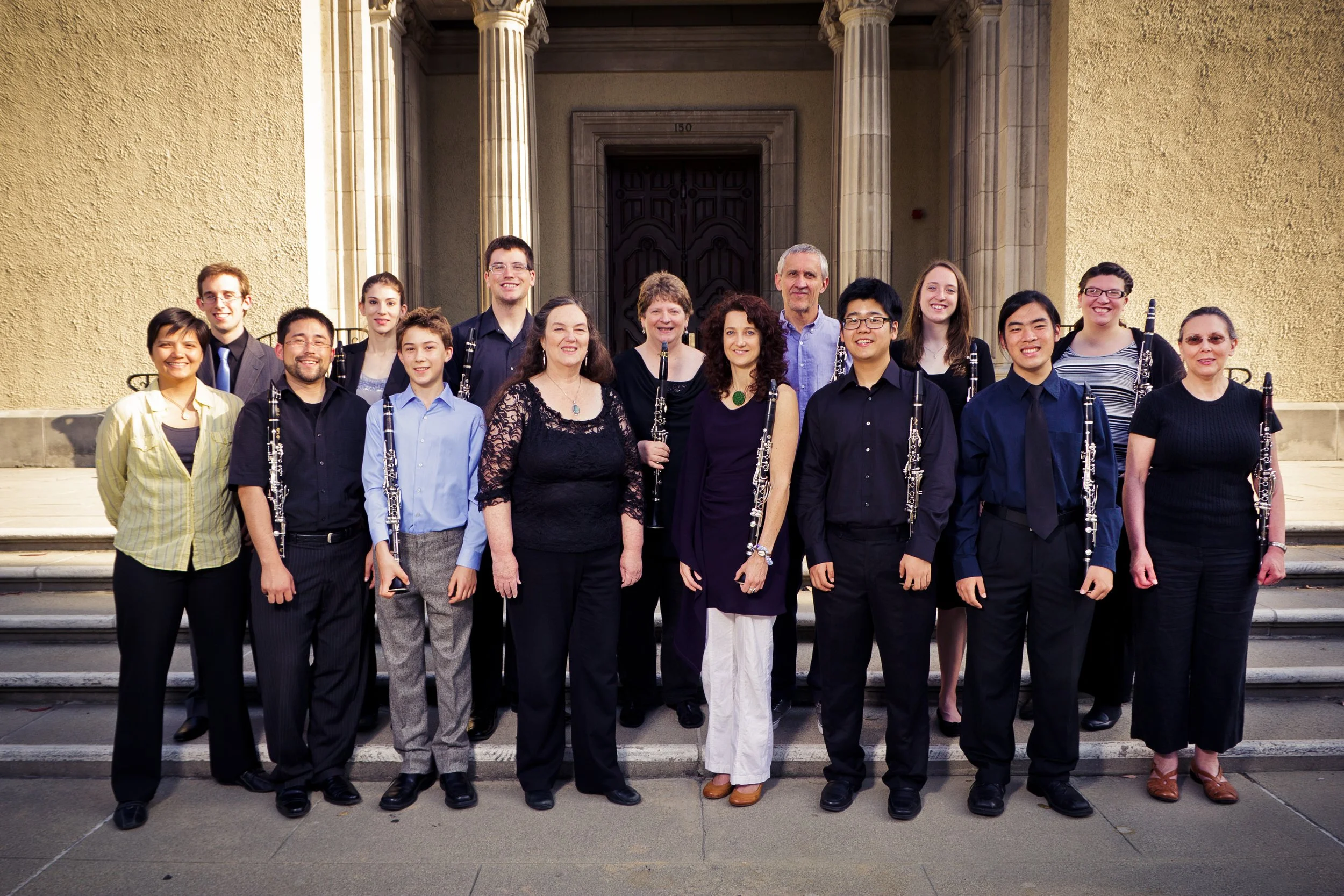 Group of people, including students and adults, standing in front of a building with stone steps, some holding clarinets, smiling for the camera.