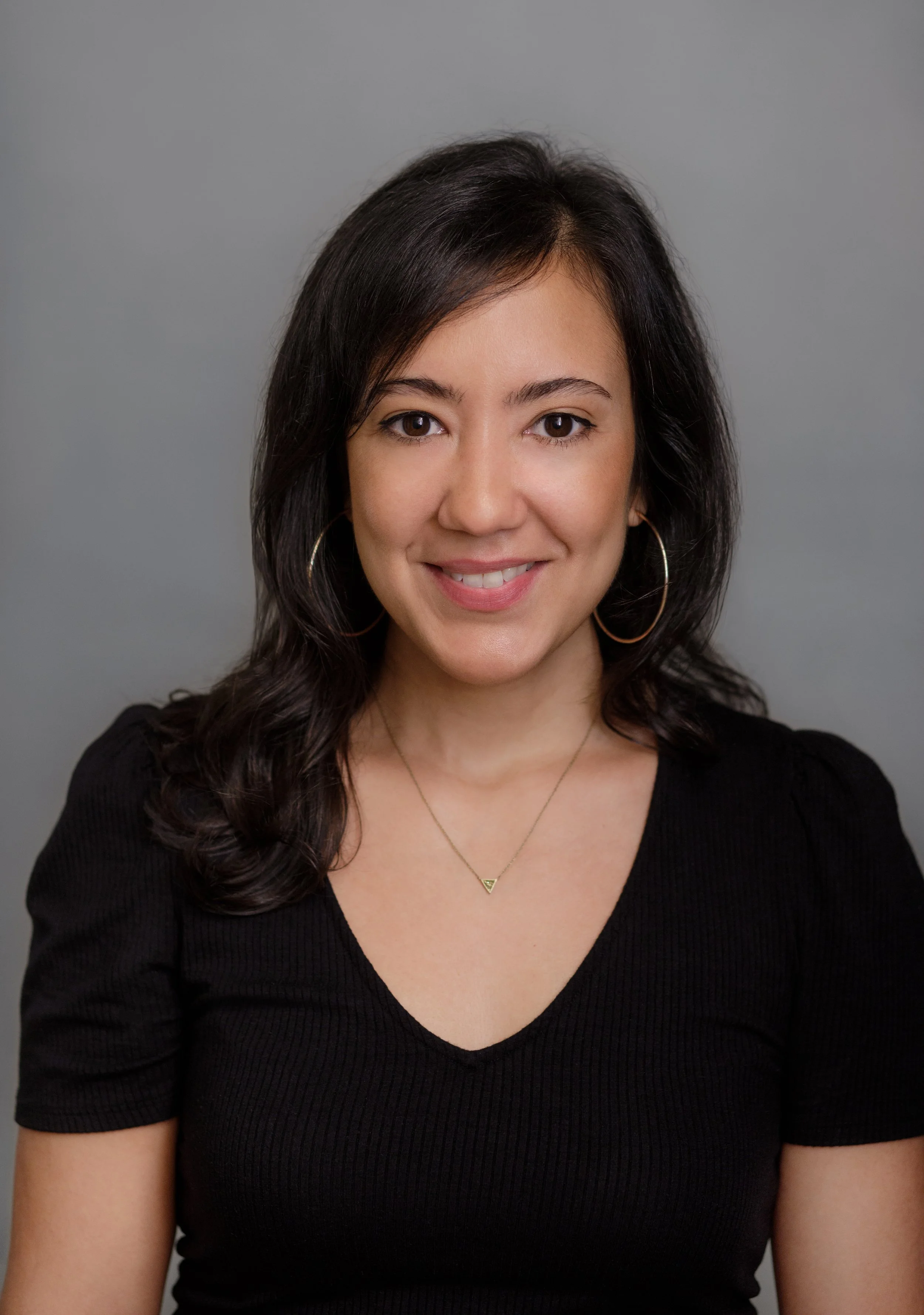Close-up portrait of a woman with dark hair, wearing hoop earrings and a black V-neck top, smiling against a gray background.