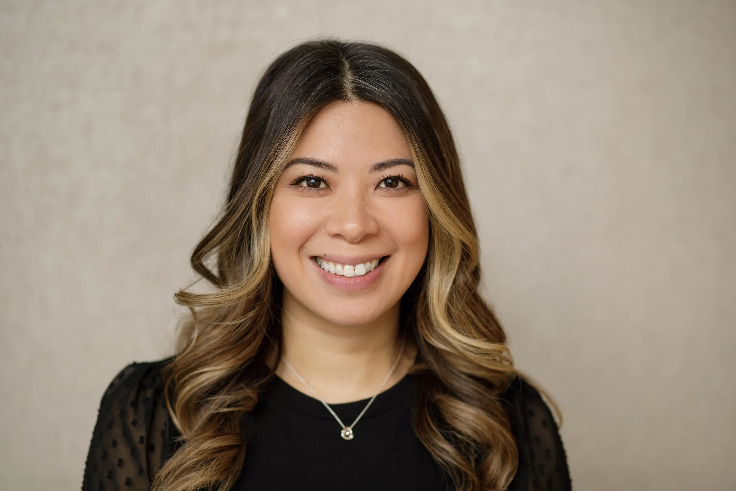 Portrait of a young woman with long, wavy hair with blonde highlights, wearing a black top and a silver necklace, smiling against a plain, neutral background.