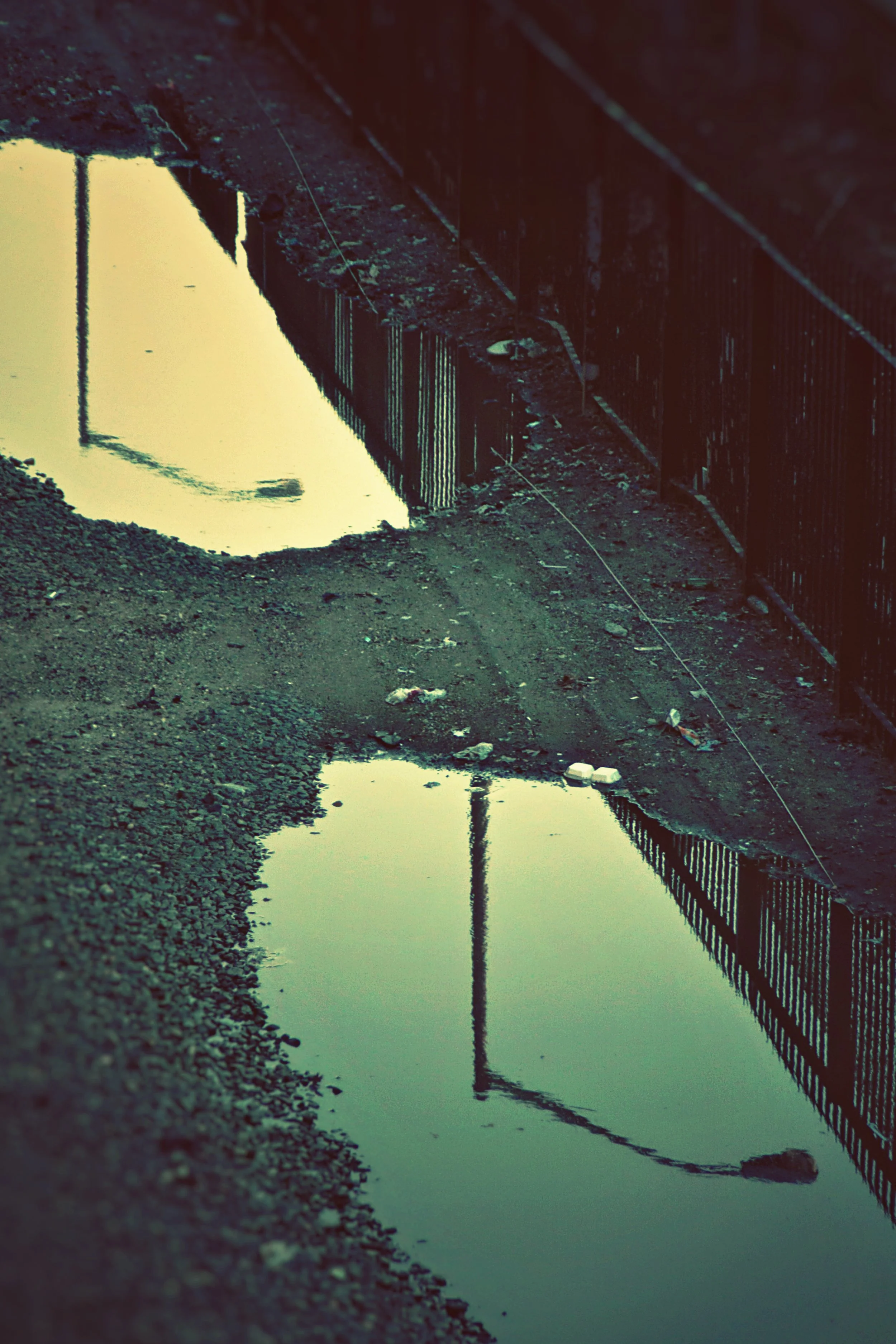 Reflection of a fence and utility pole in muddy water with gravel along the edge.