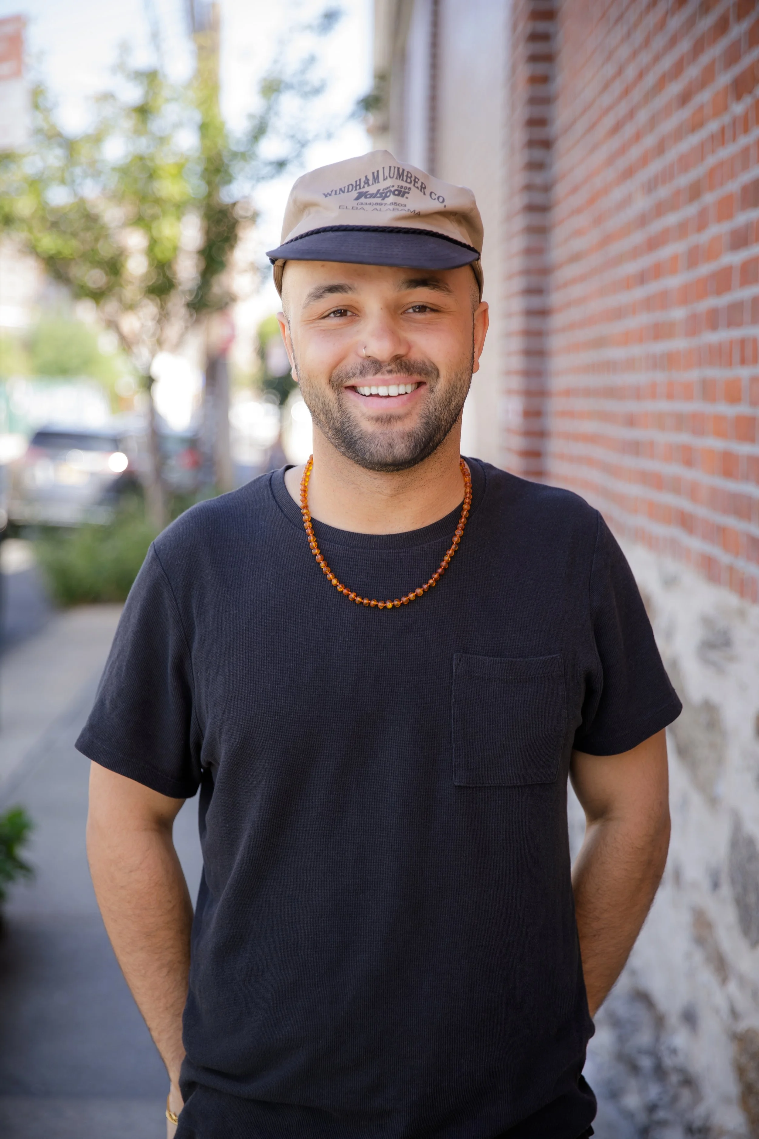 Smiling man wearing a black t-shirt, an amber beaded necklace, and a beige cap with black brim, standing in front of a brick wall and blurred trees.