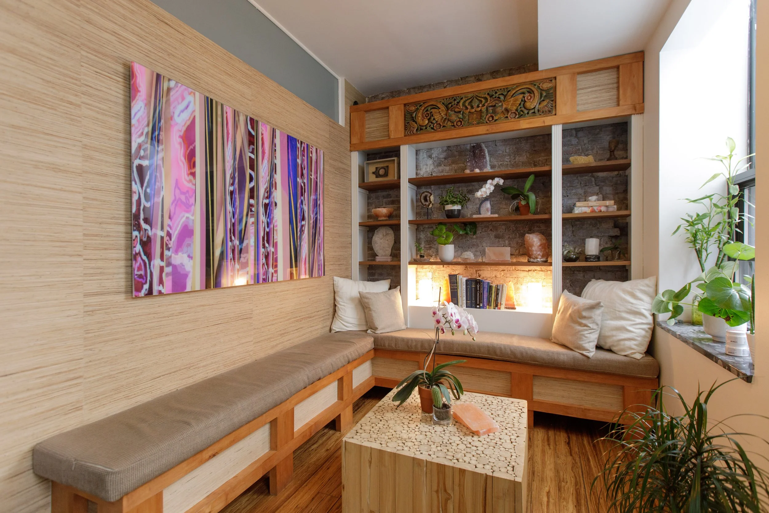 Cozy seating nook with built-in bench with beige cushions, surrounding a small table with potted plants, and decorated with a colorful abstract painting, wooden shelves with plants and decorative objects, and a textured brick wall in the background, 