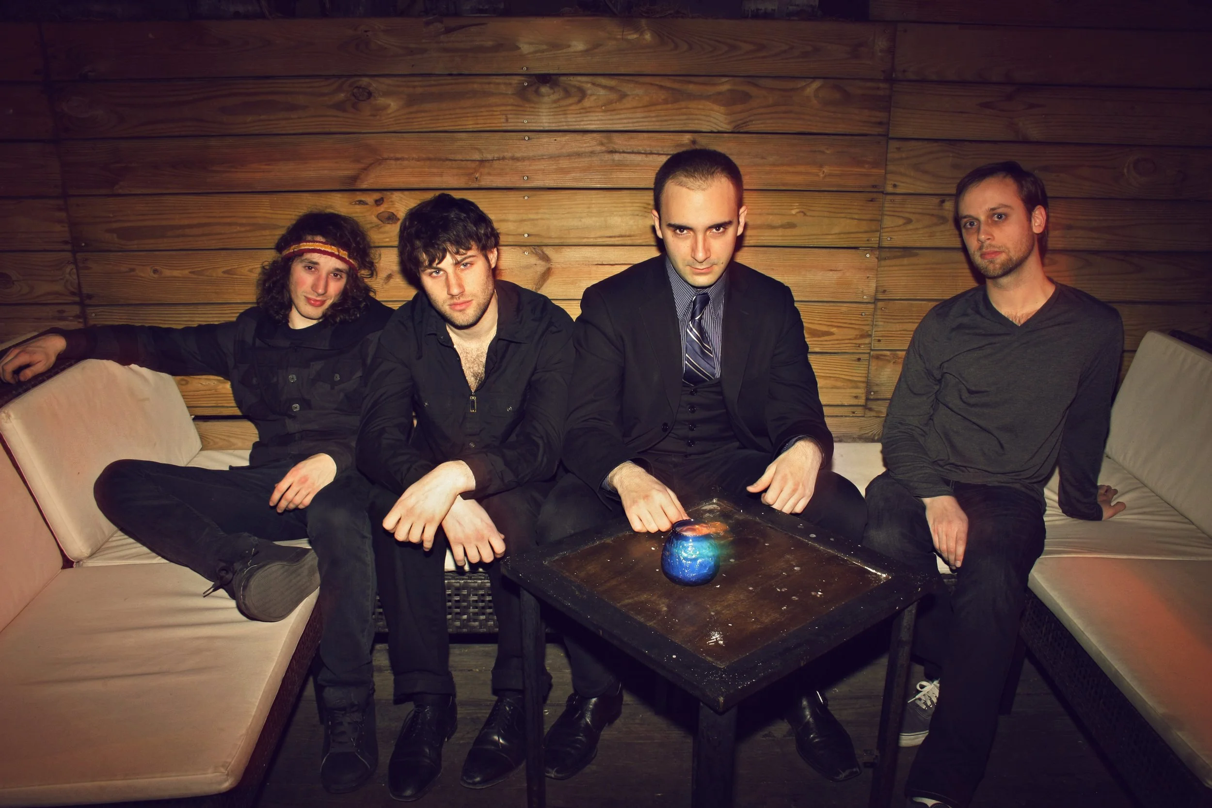 Four young men sitting on a beige couch and a bench inside a wooden-paneled room, with a dark rectangular table in front of them, holding a small blue and orange decorative object.