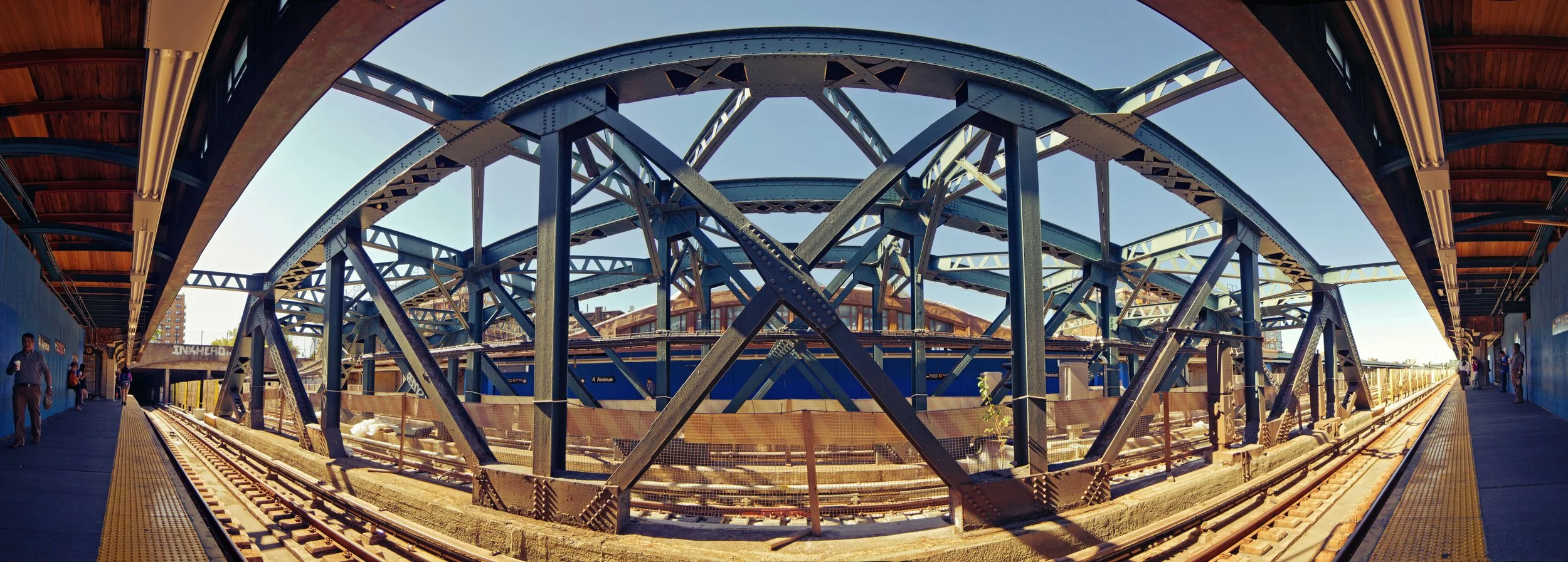View of a train station with an elevated wooden platform on the sides and a large blue steel structure supporting the tracks and overhead rail system in the center. Several people are walking on the platform. Clear blue sky is visible above.