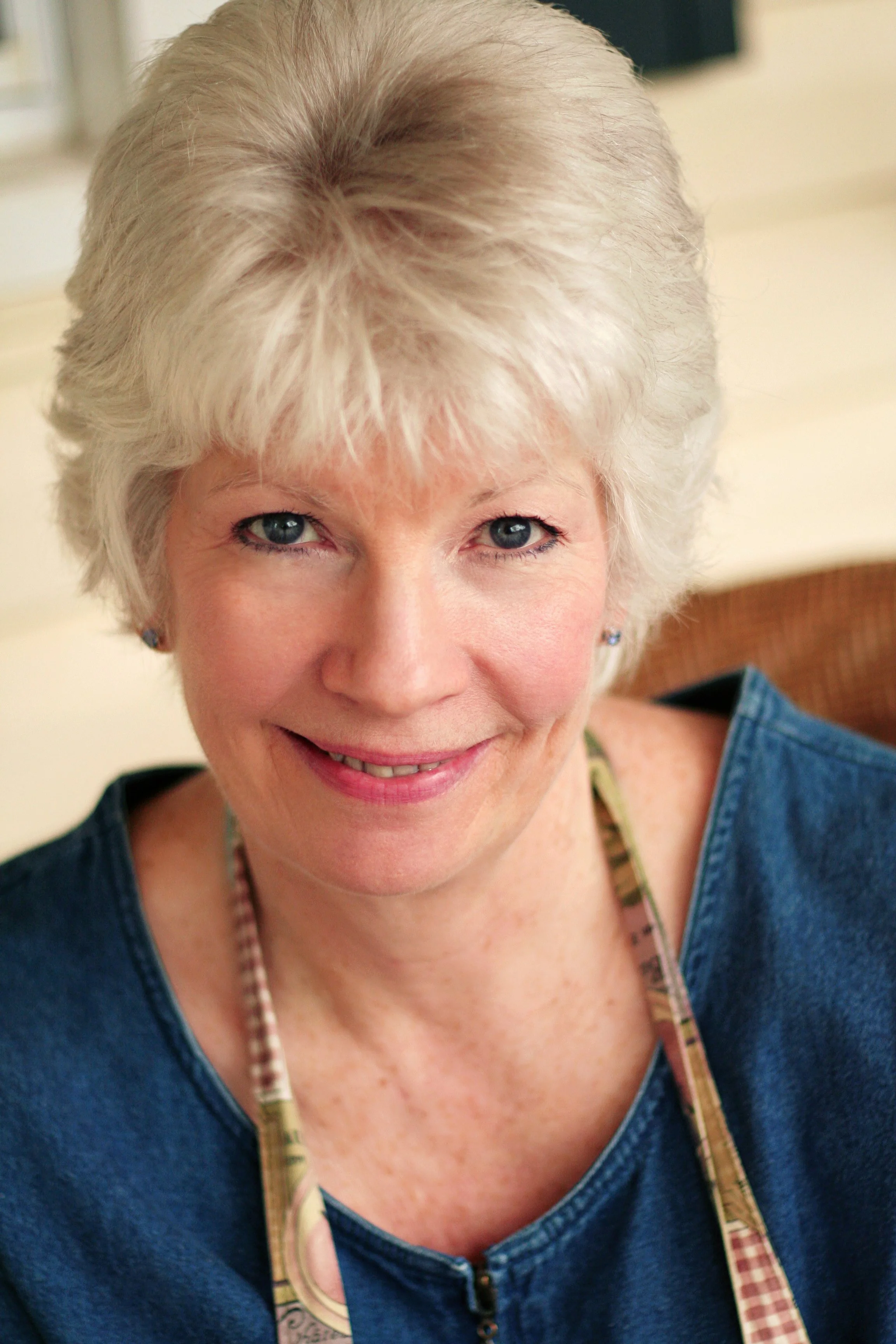 Close-up of an older woman with short, curly, gray hair, blue eyes, and light skin, smiling and wearing a blue shirt, an apron, and a beaded necklace.