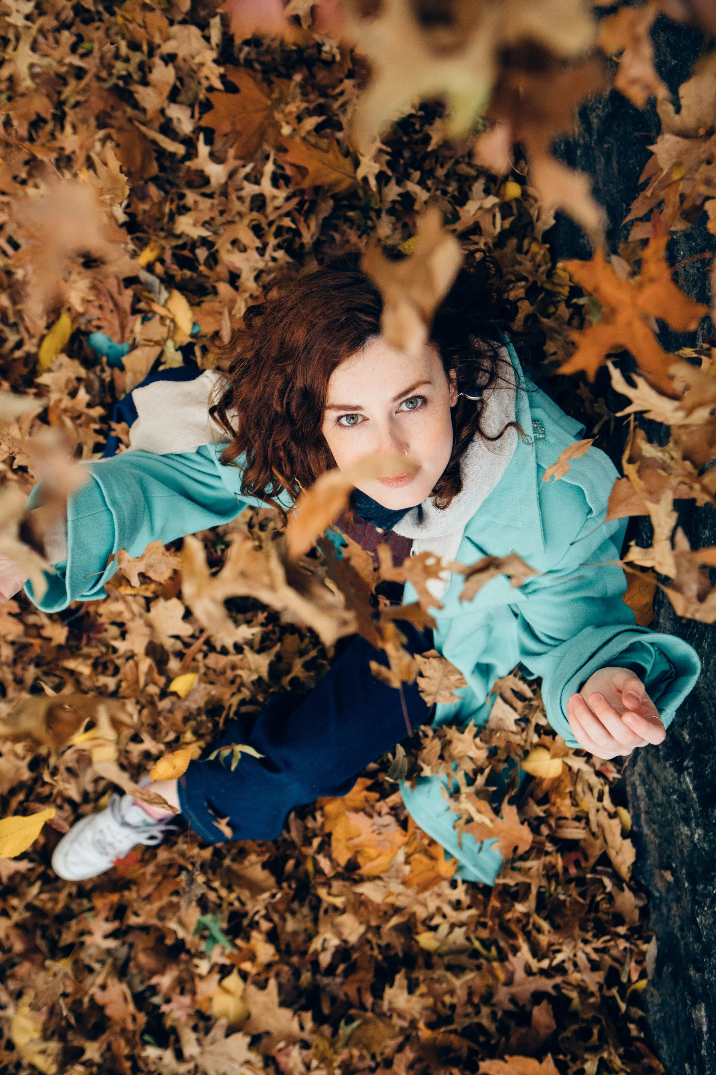A young woman with curly brown hair lying on her back in a pile of fall leaves, looking up at the camera, wearing a teal jacket and white sneakers.