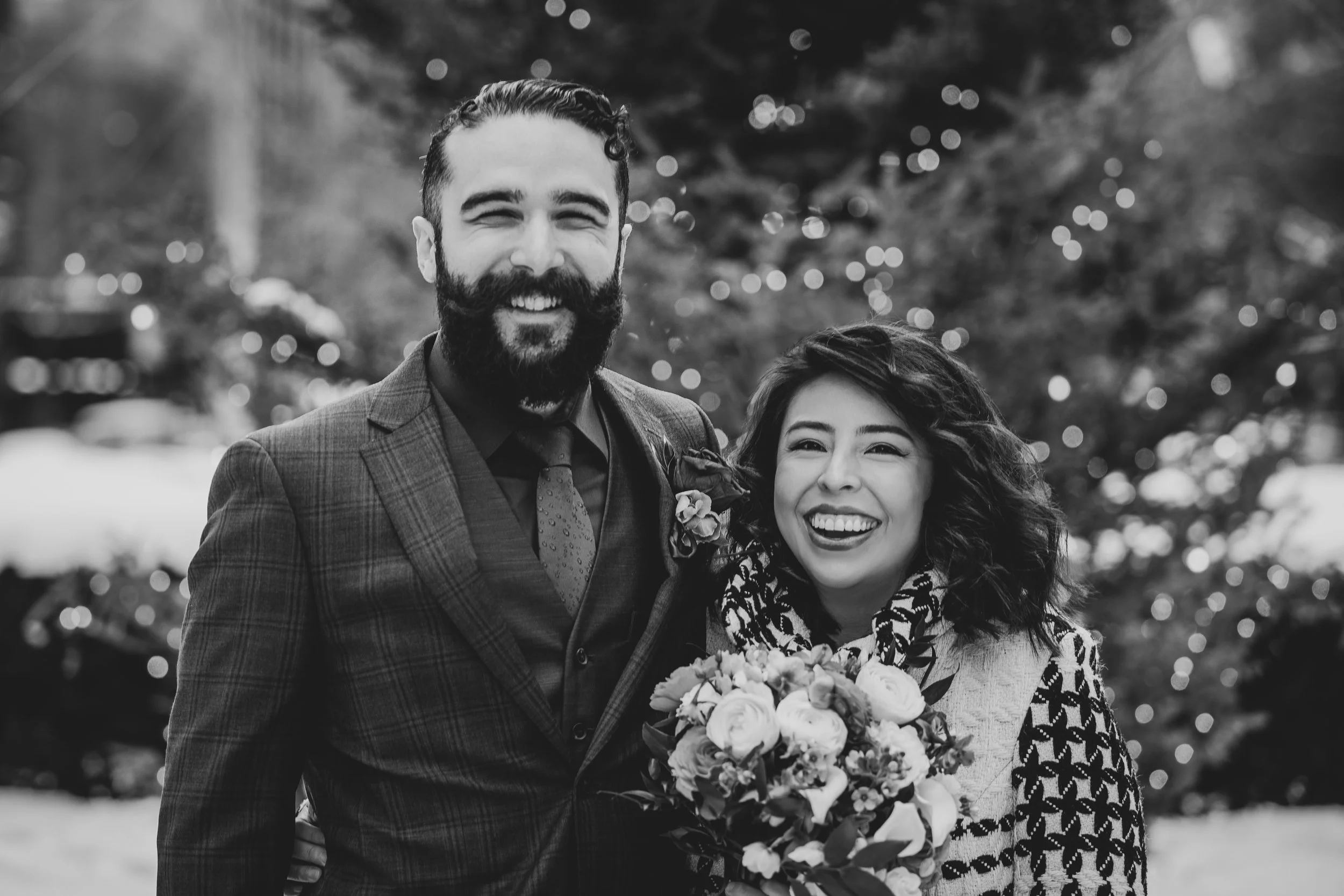 A smiling man with a beard and a woman with curly hair holding a bouquet of flowers outdoors, with bokeh lights in the background.