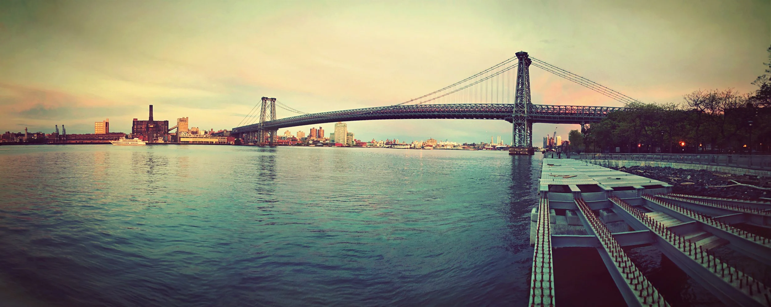 View of a large steel suspension bridge spanning a wide river, with city buildings in the background and trees on the right side; water in the foreground reflects the soft evening sky.