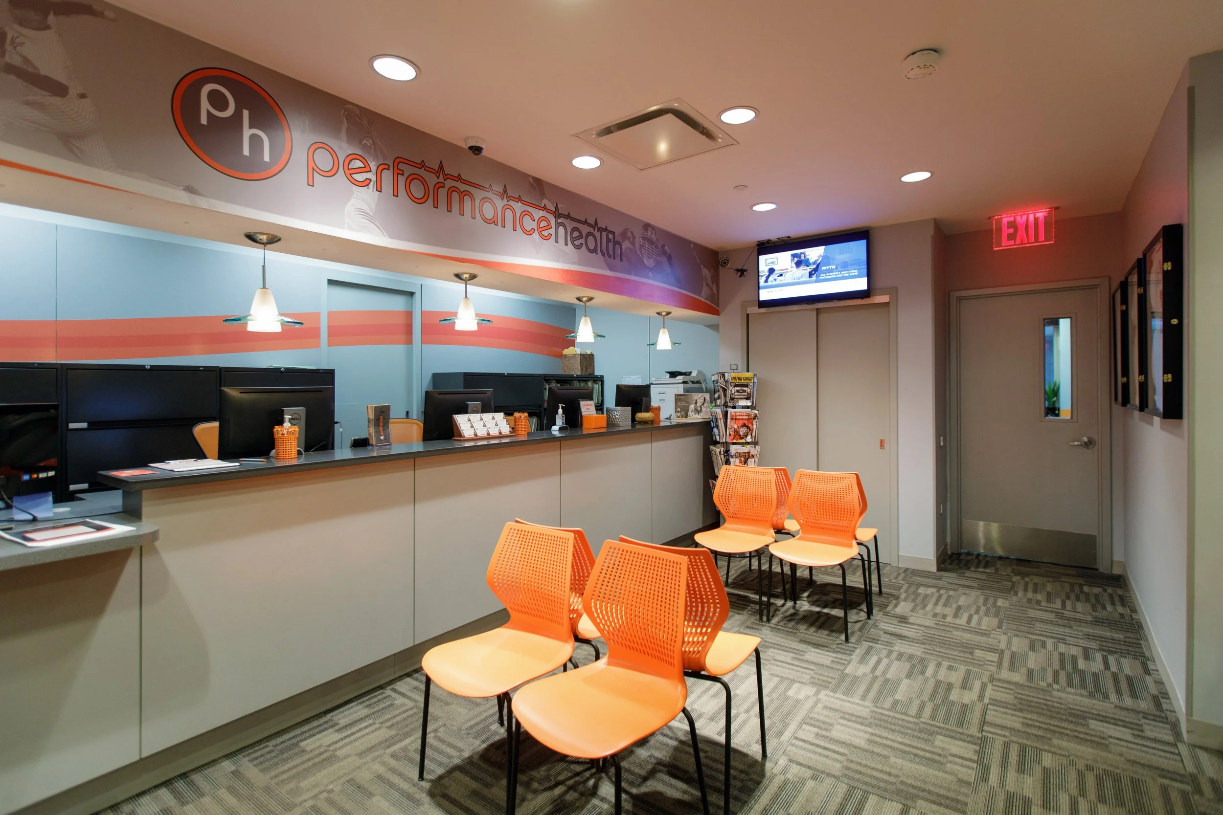 A hospital or clinic reception area with a counter, computers, and orange chairs. The wall behind the counter has a sign that reads 'performance health,' and there is a digital screen and exit sign visible.