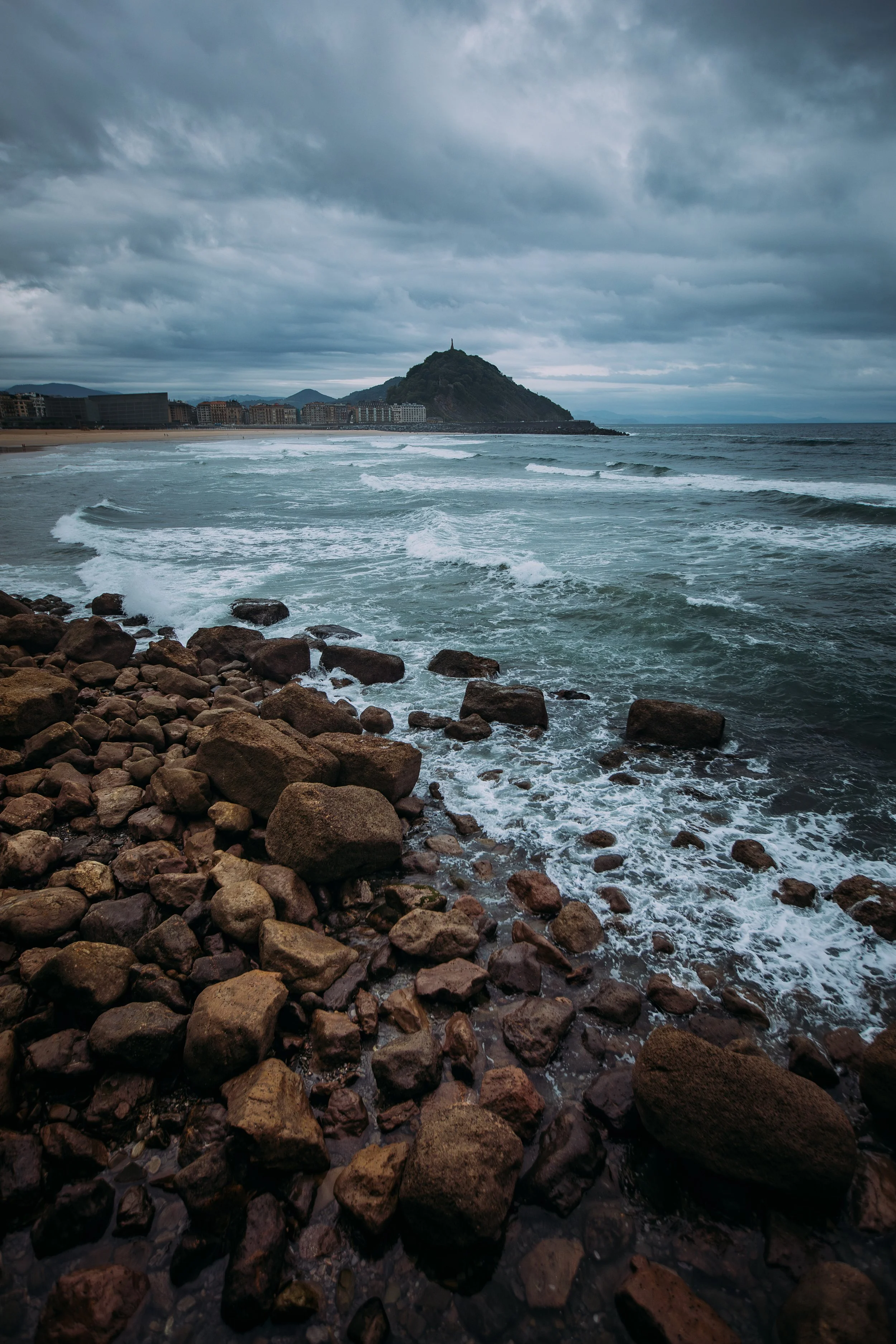 A rocky shoreline with waves crashing against the rocks, a distant cityscape with buildings near the coast, and a hill with a statue on top under a cloudy sky.