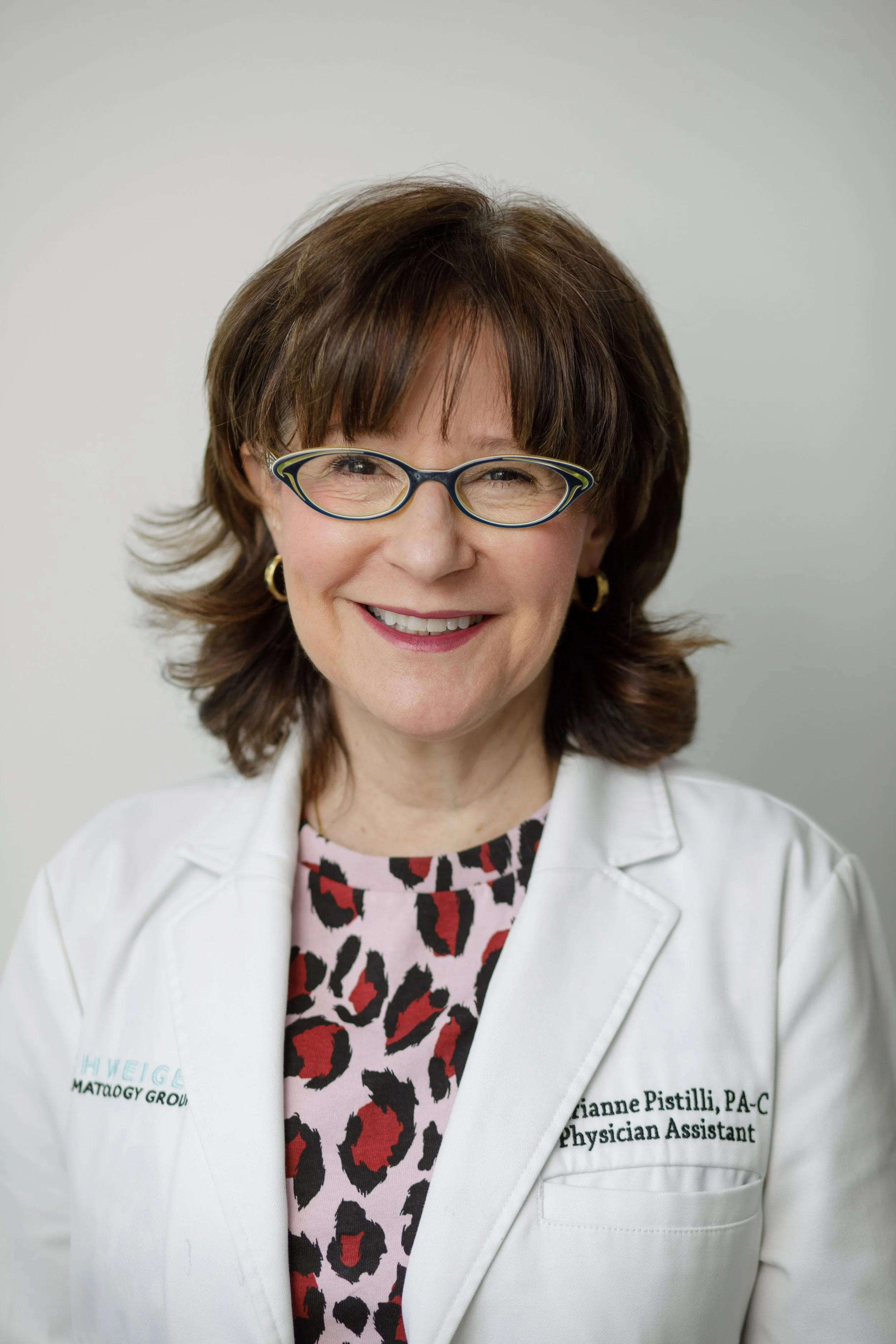 Smiling woman with short brown hair, wearing glasses, gold earrings, a white lab coat with embroidery, and a pink leopard print top, against a plain gray background.