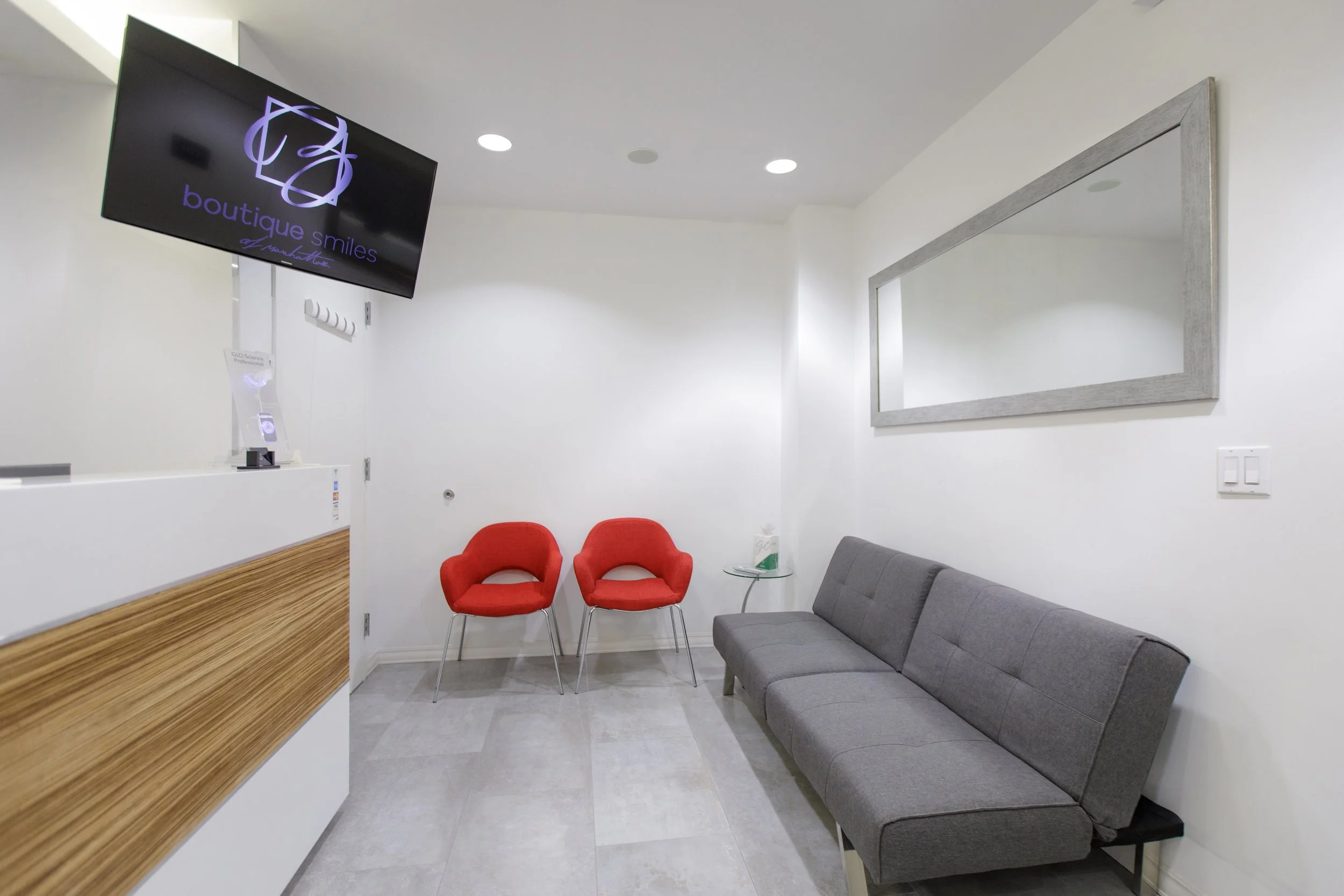 Waiting room with a gray sofa, two red chairs, a small glass side table, a large mirror, and an overhead TV displaying 'boutique smiles.'
