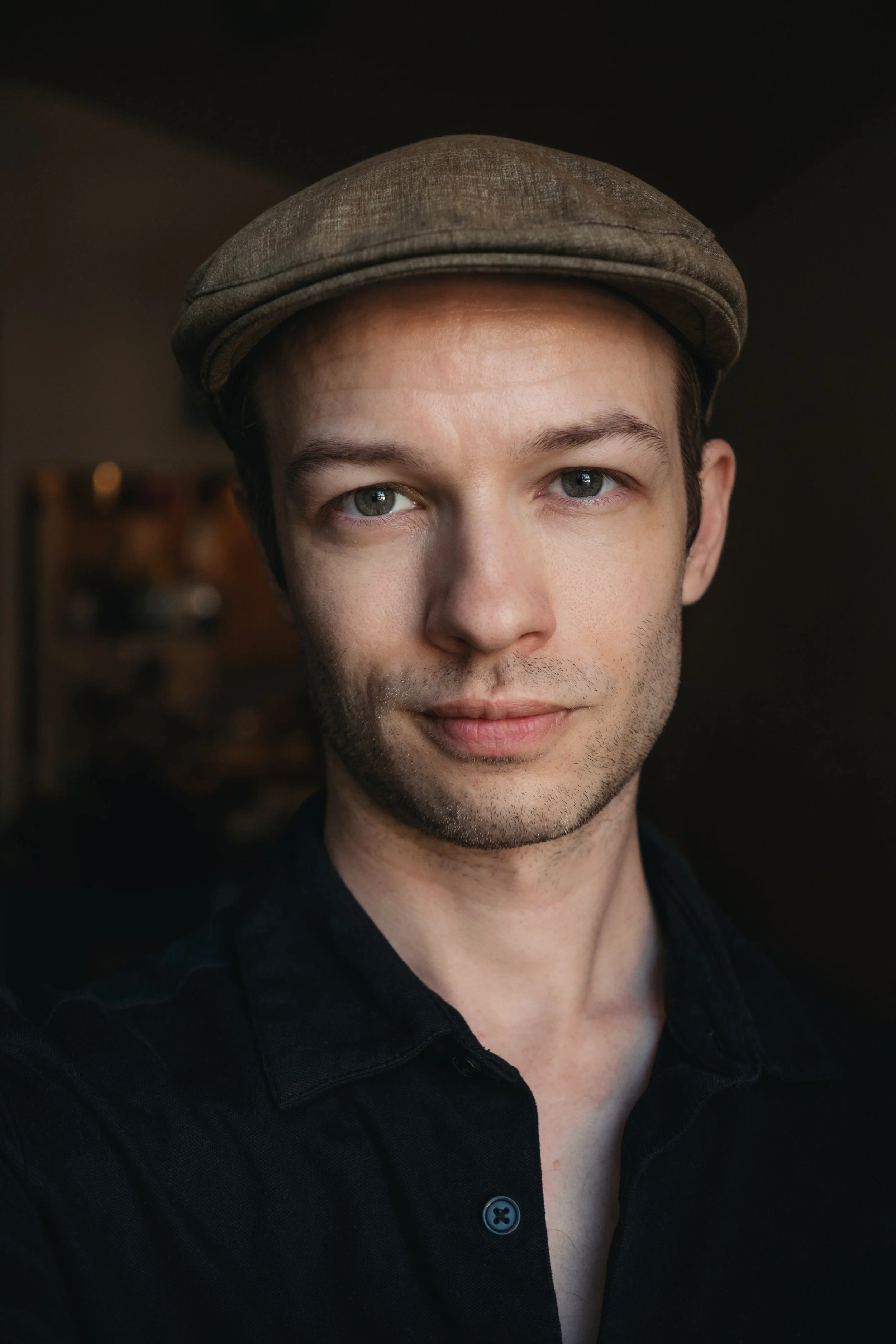 A young man wearing a brown flat cap and a black shirt, with blue eyes and a light beard, smiling slightly, indoors with a blurred background.
